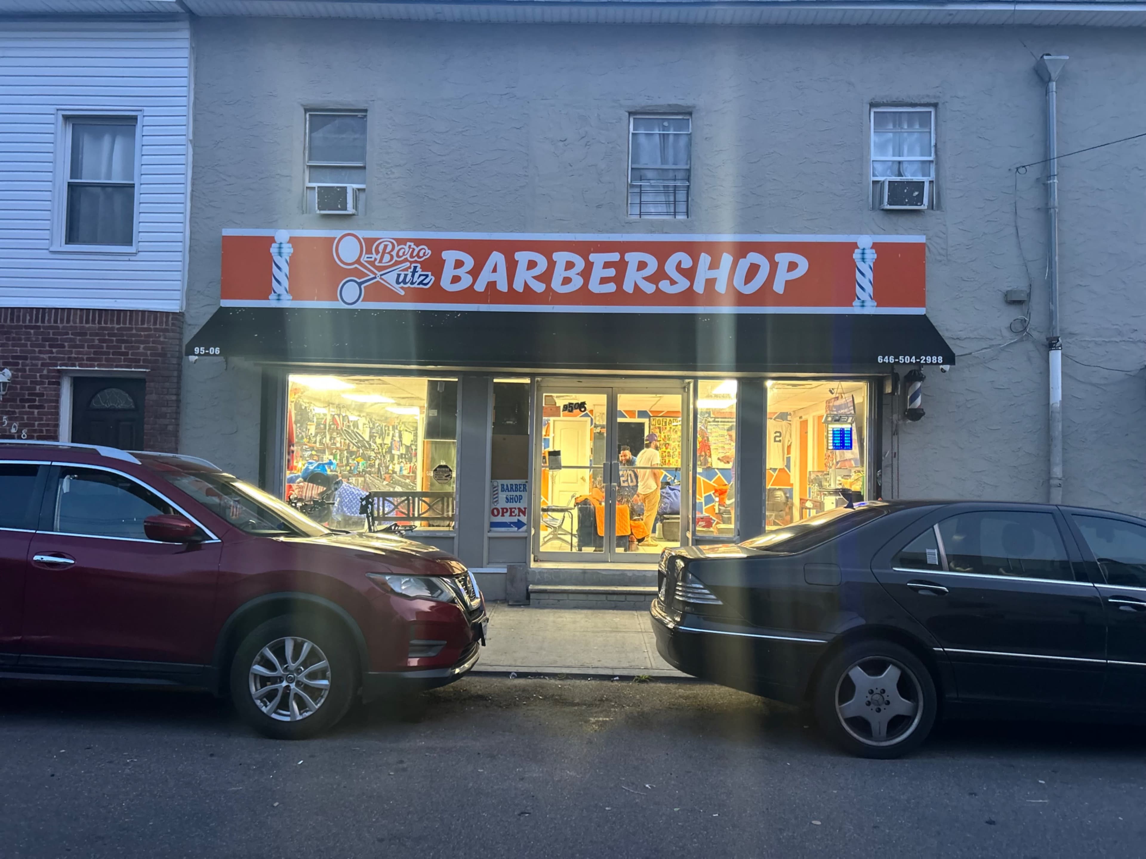 The image shows a barbershop with a bright sign reading "Barbershop," located between two parked cars on a street.