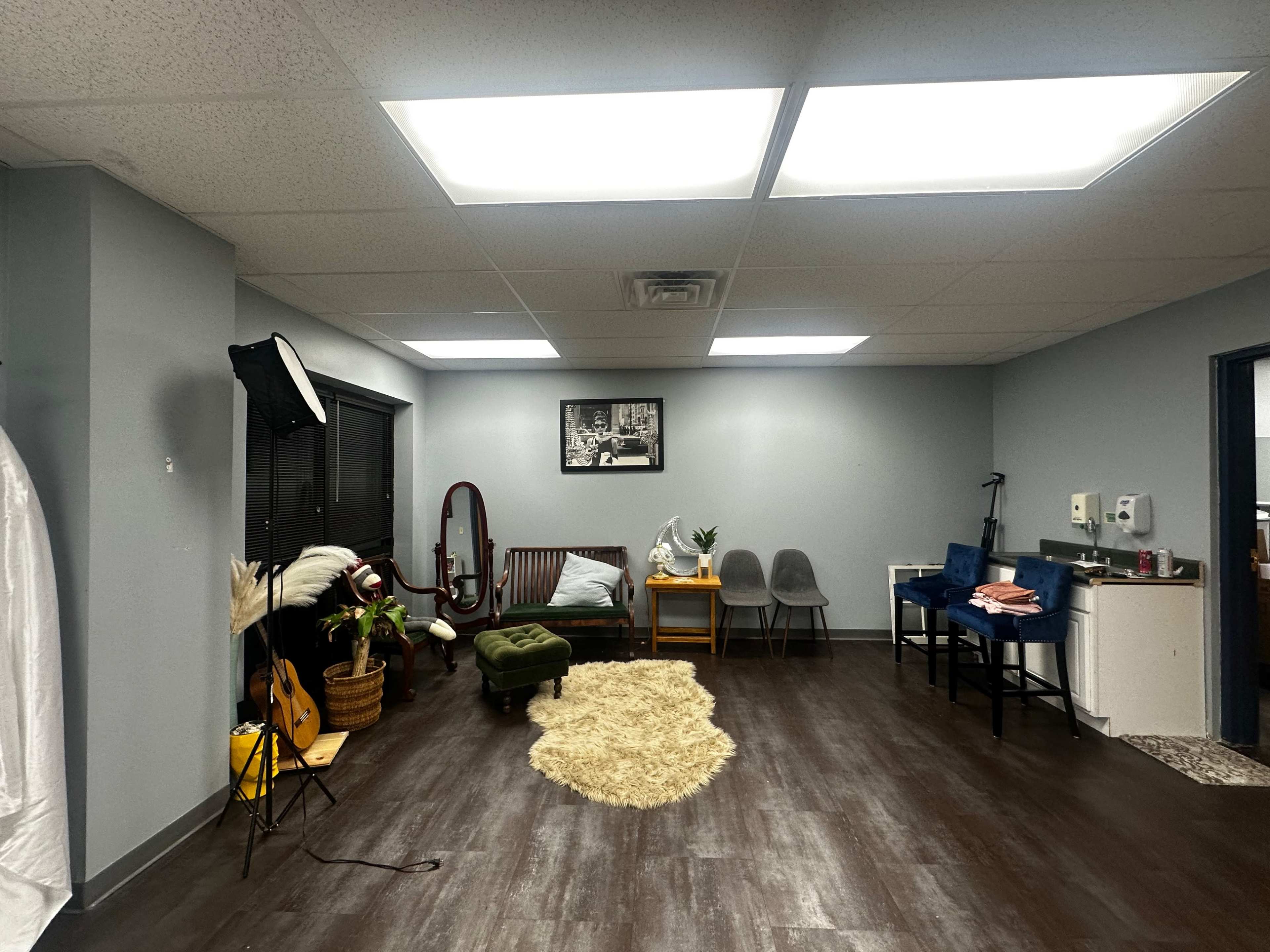 The image shows a spacious room with gray walls, a hardwood floor, a seating area that includes a couch and chairs, and various decorations like a guitar, plants, and a framed picture on the wall.