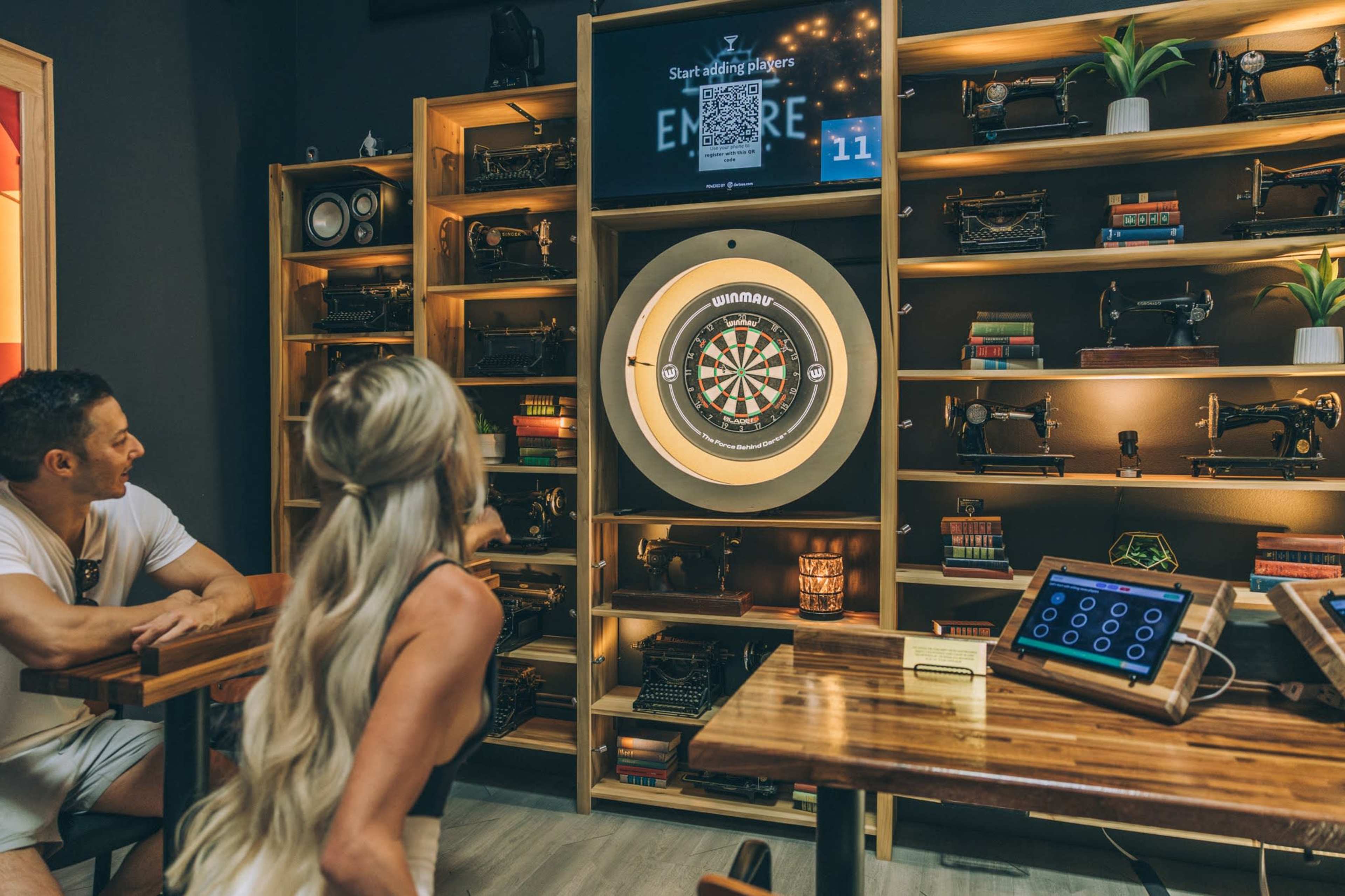 A woman prepares to throw a dart while a man observes in a modern game room featuring a dartboard and decorative shelves with various vintage items.
