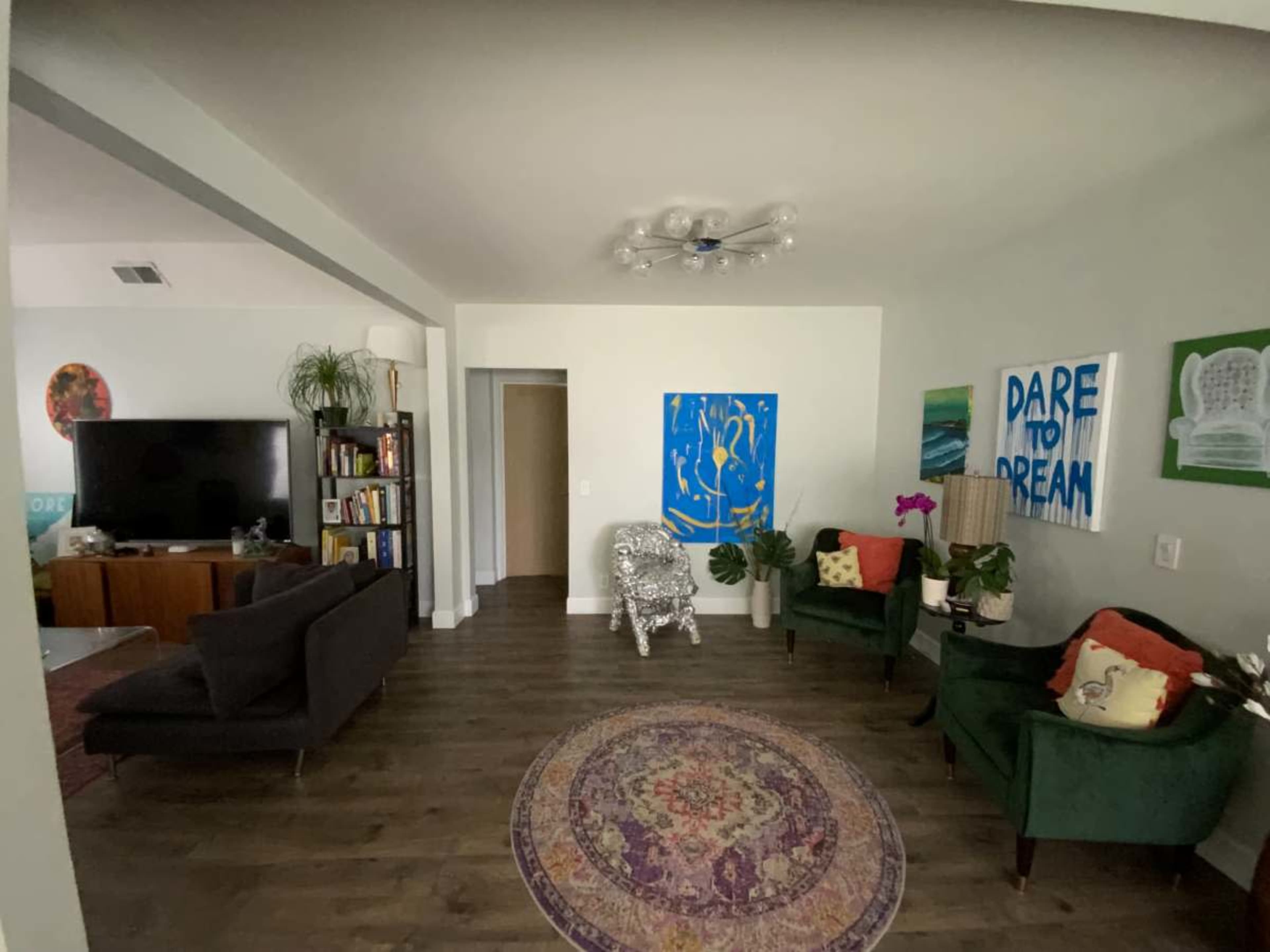 The image shows a living room with a television, bookshelves, colorful art on the walls, and green seating arranged around a patterned rug.