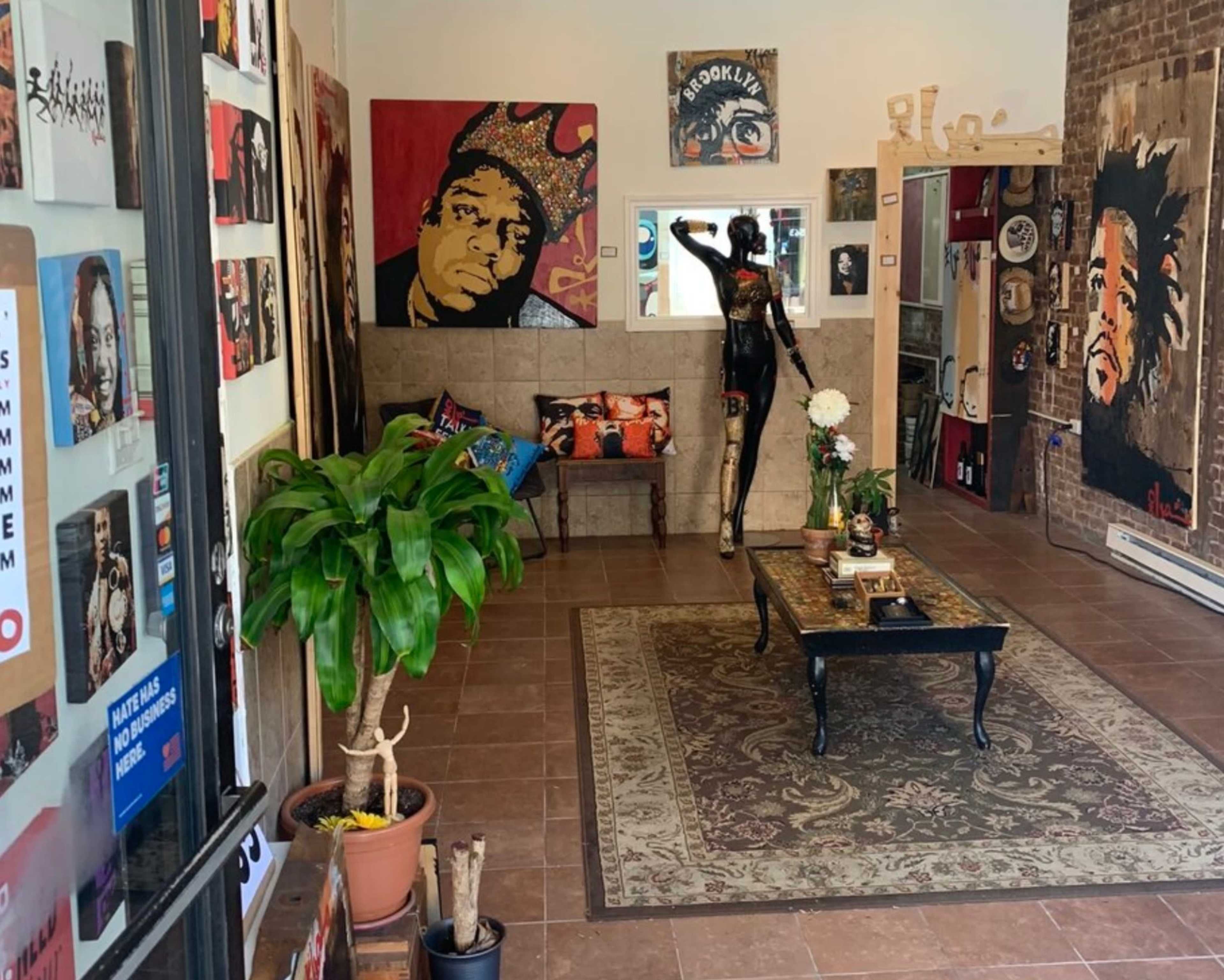 An art gallery featuring vibrant portraits on the walls, a coffee table in the center, and a mannequin dressed in black, surrounded by decorative plants and furnishings.