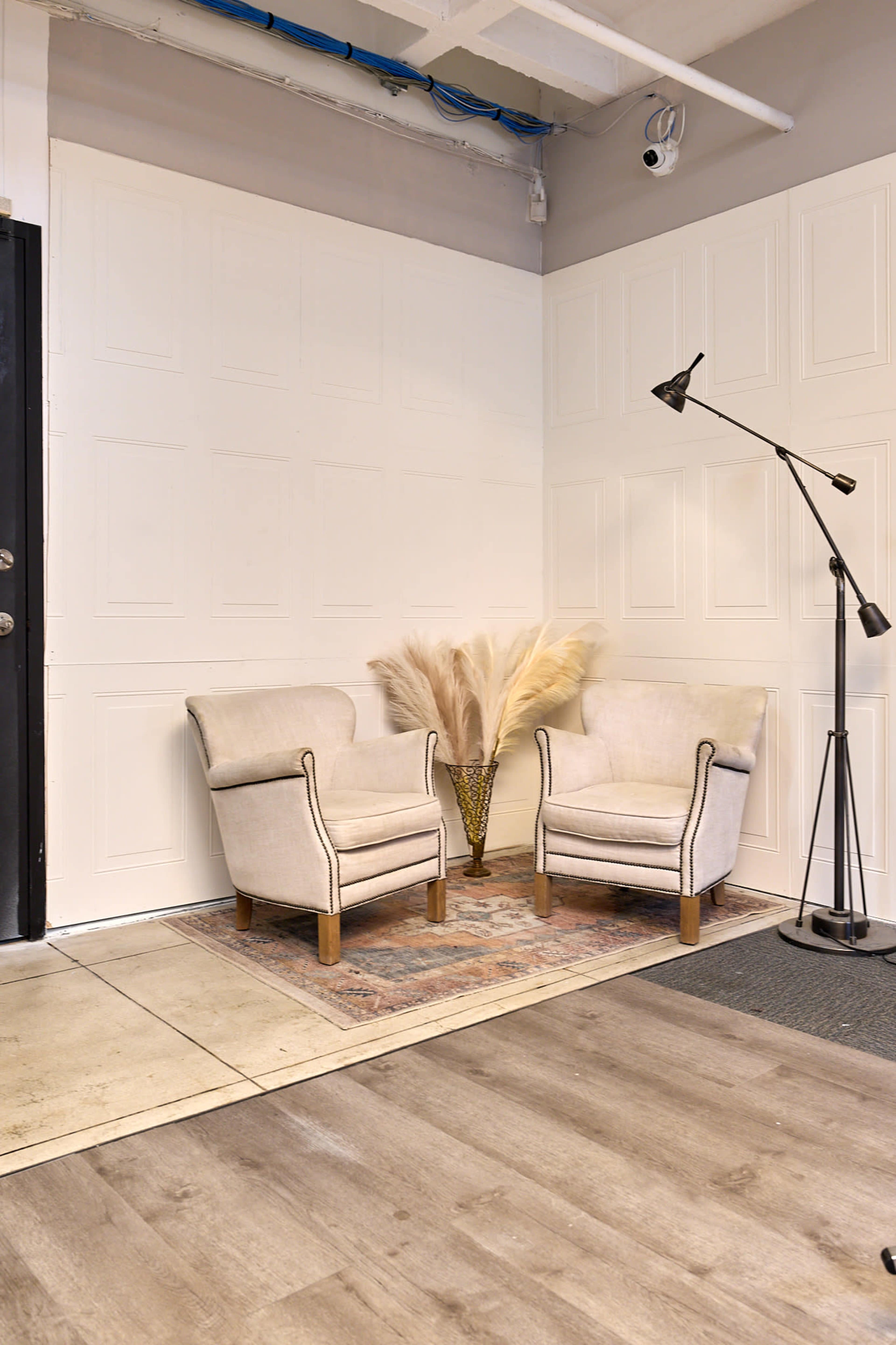 The image shows a small room with two light-colored armchairs positioned on a patterned rug, accompanied by a floor lamp and a decorative vase with dried plants.