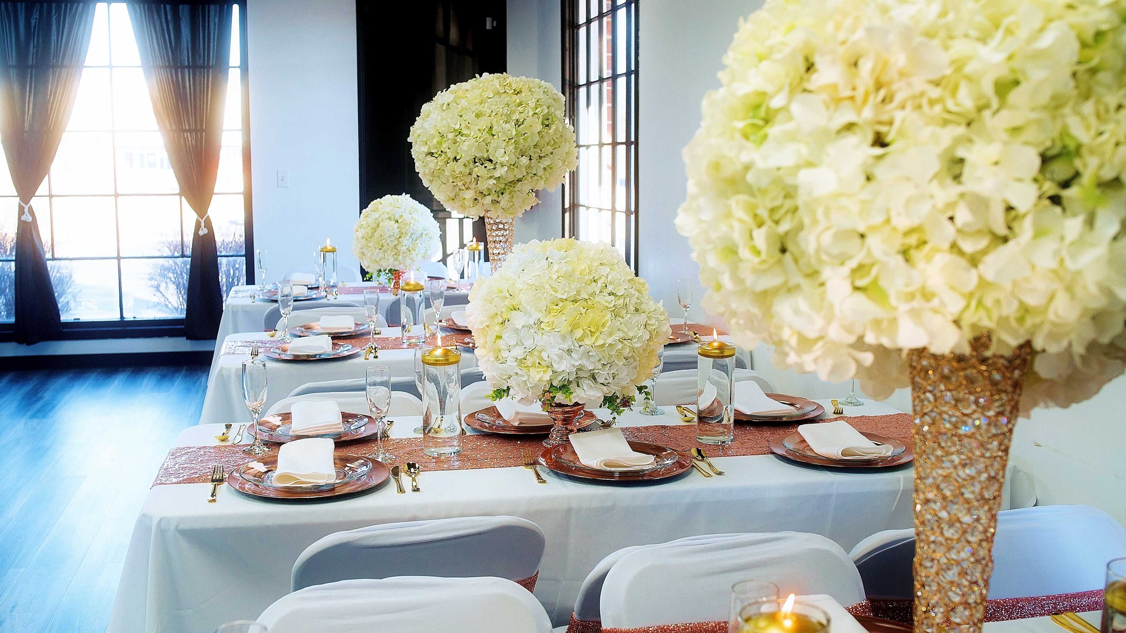 A elegantly set dining area with tables adorned with white floral centerpieces and sparkling table runners, illuminated by natural light through large windows.