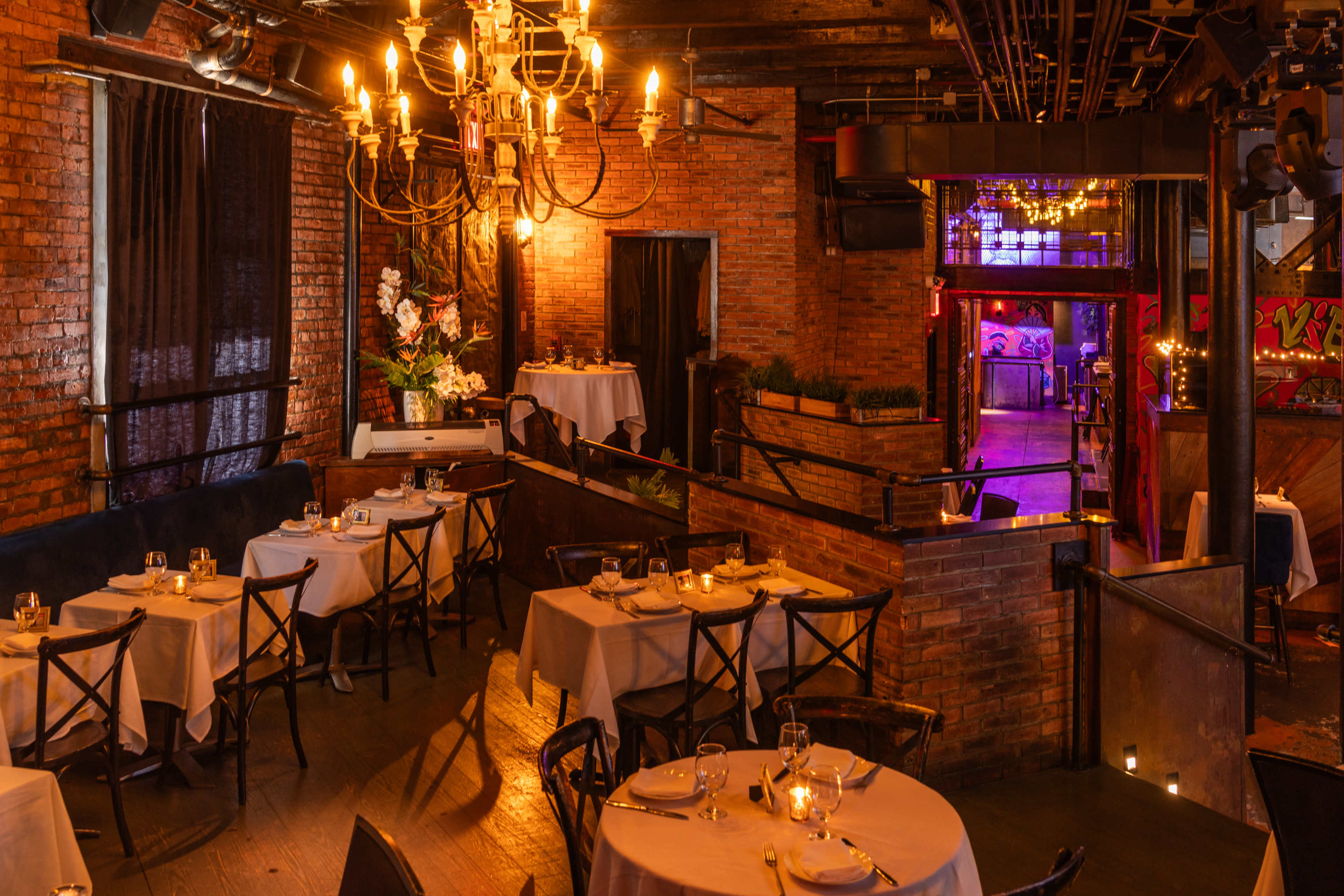 The image shows an upscale restaurant with wooden floors, exposed brick walls, and tables set for dining under a chandelier.