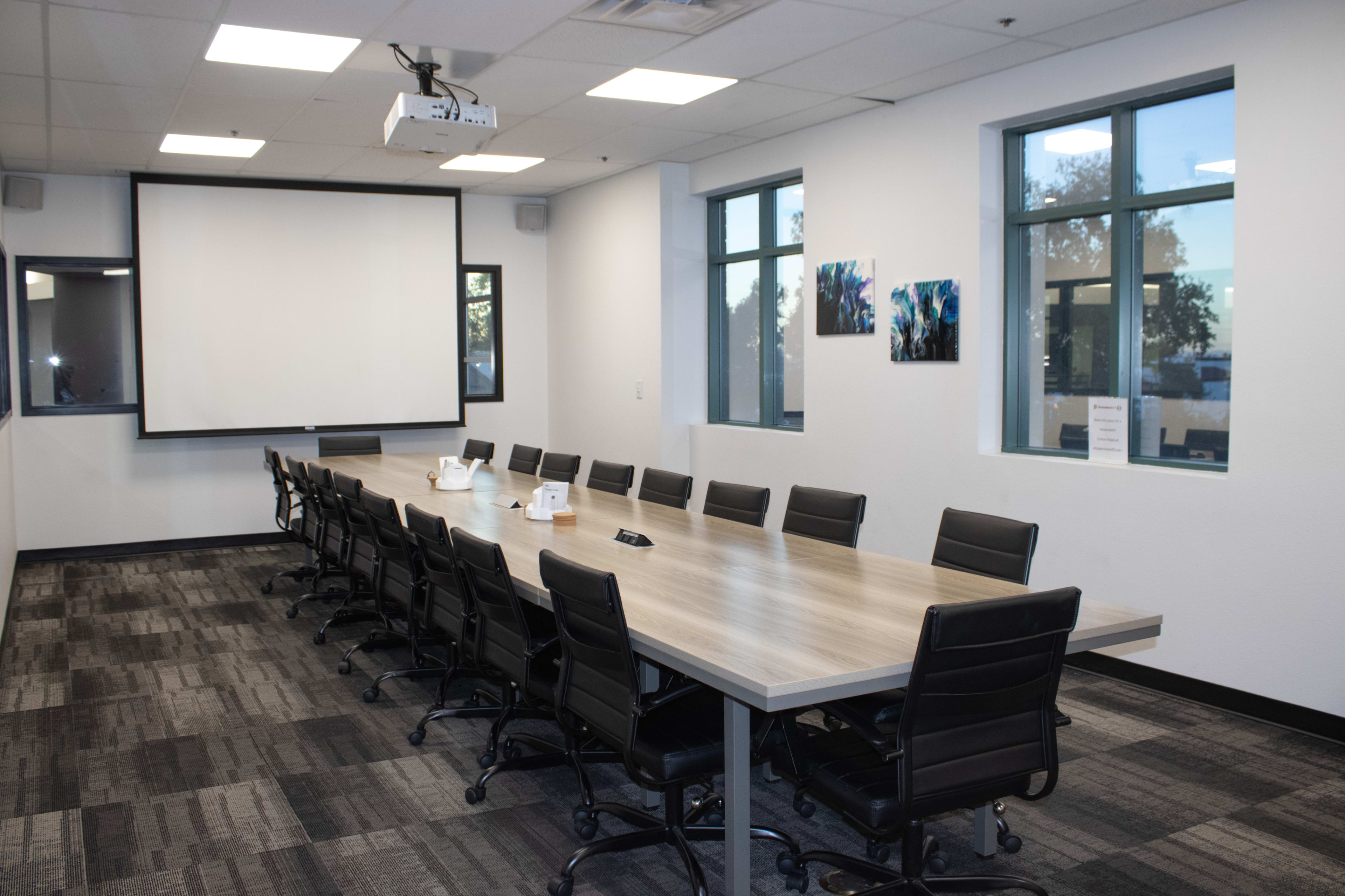 A long conference table with black chairs is set up in a well-lit room, featuring large windows and a projector screen at one end.