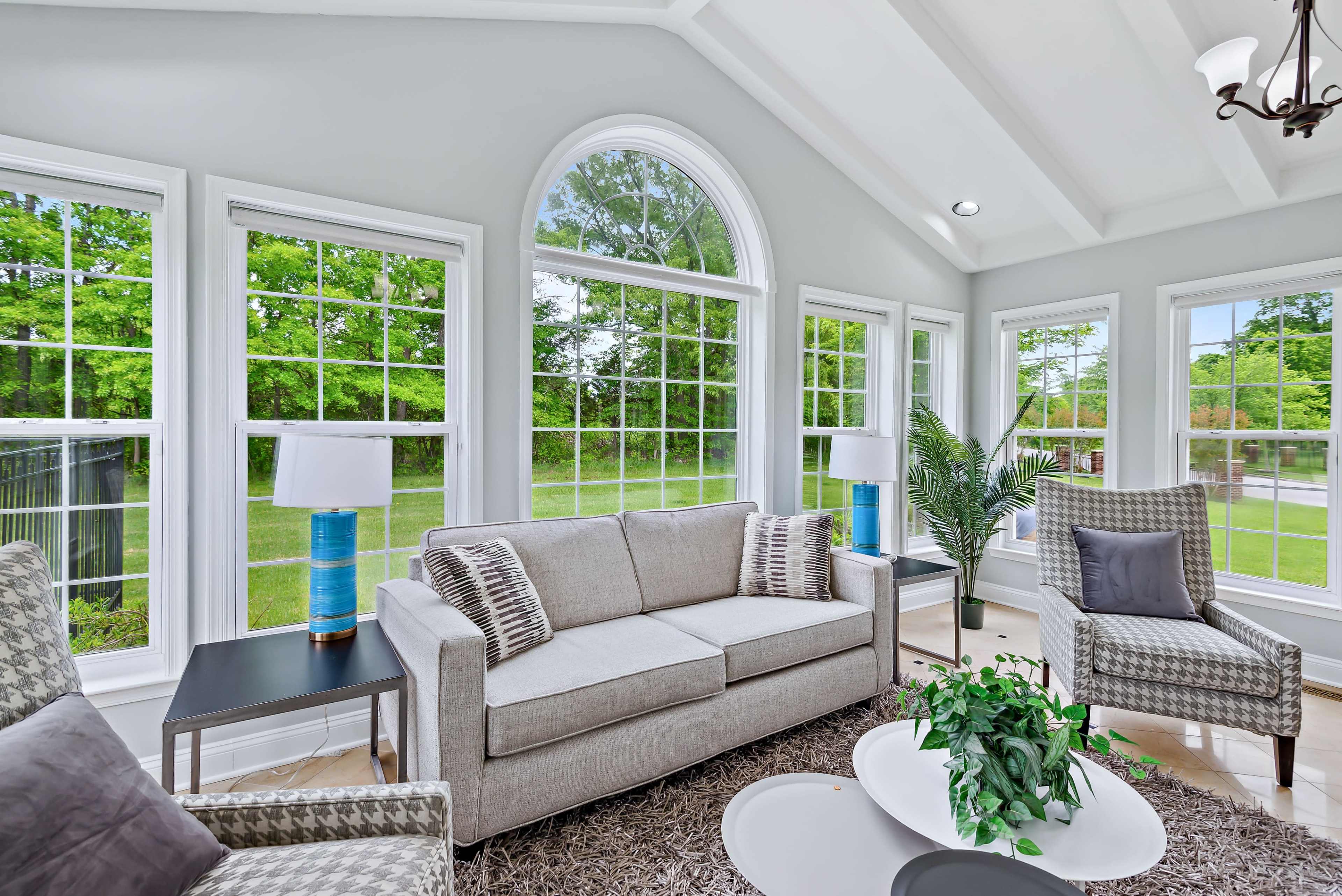 The image shows a bright sunroom featuring a gray sofa, two armchairs, a coffee table, and large windows that overlook a green landscape.