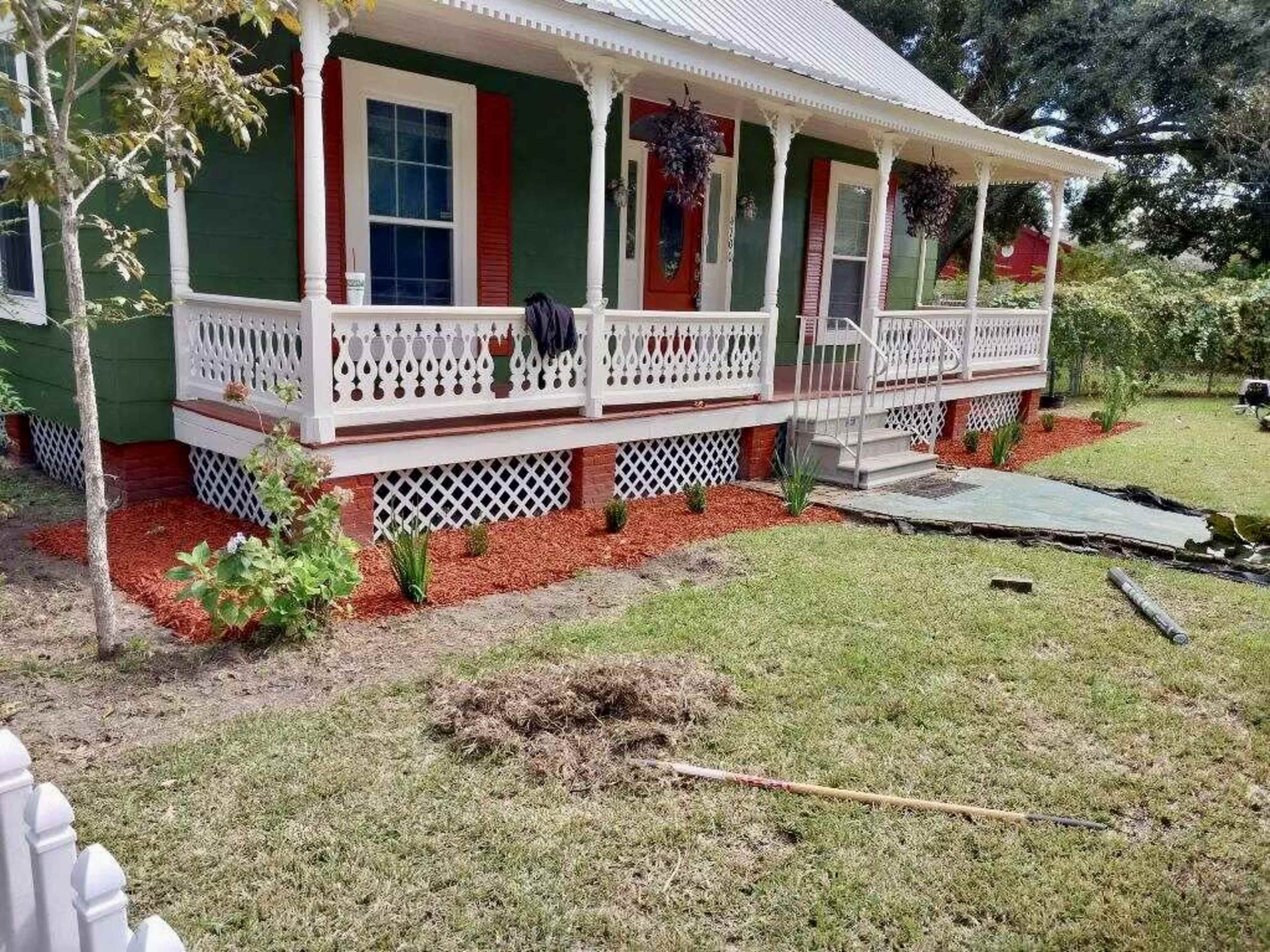 A colorful house with green siding, white trim, and a porch is surrounded by freshly mulched flower beds and newly planted greenery.