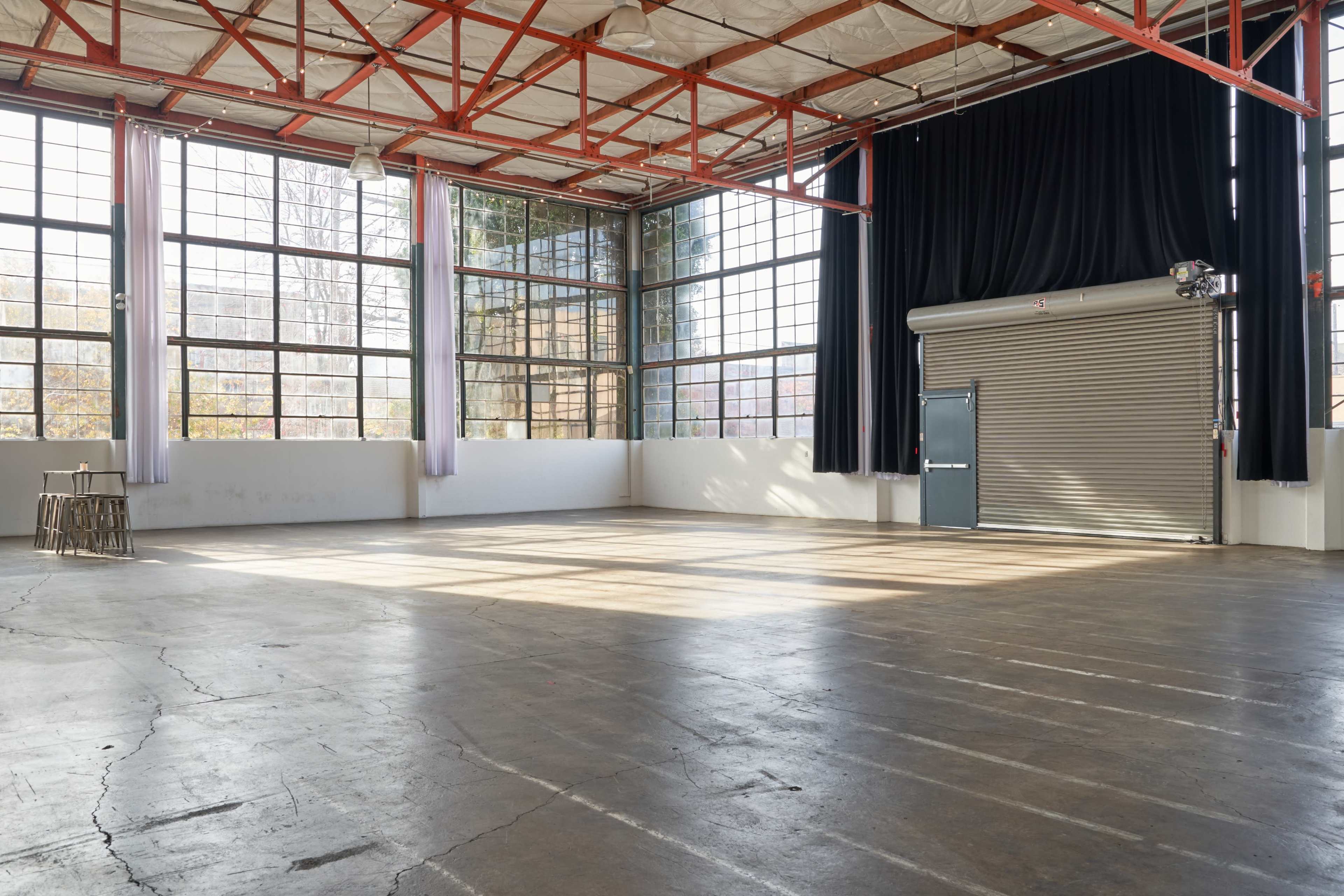 A spacious industrial room with large windows, concrete flooring, and a rolled-up door at one end.