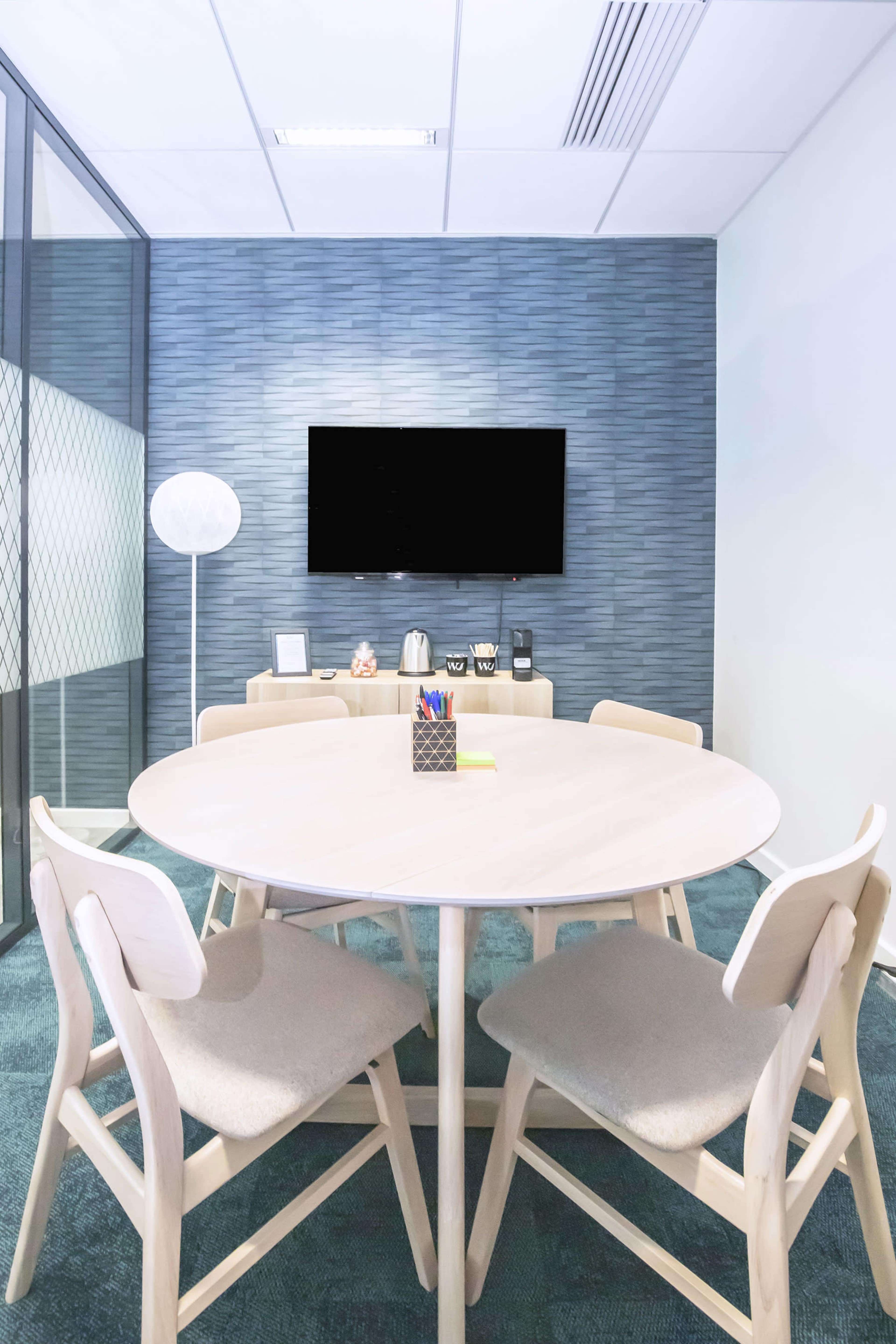 A round wooden table surrounded by four chairs is set in a modern conference room with a television mounted on the wall and a small kitchenette in the background.