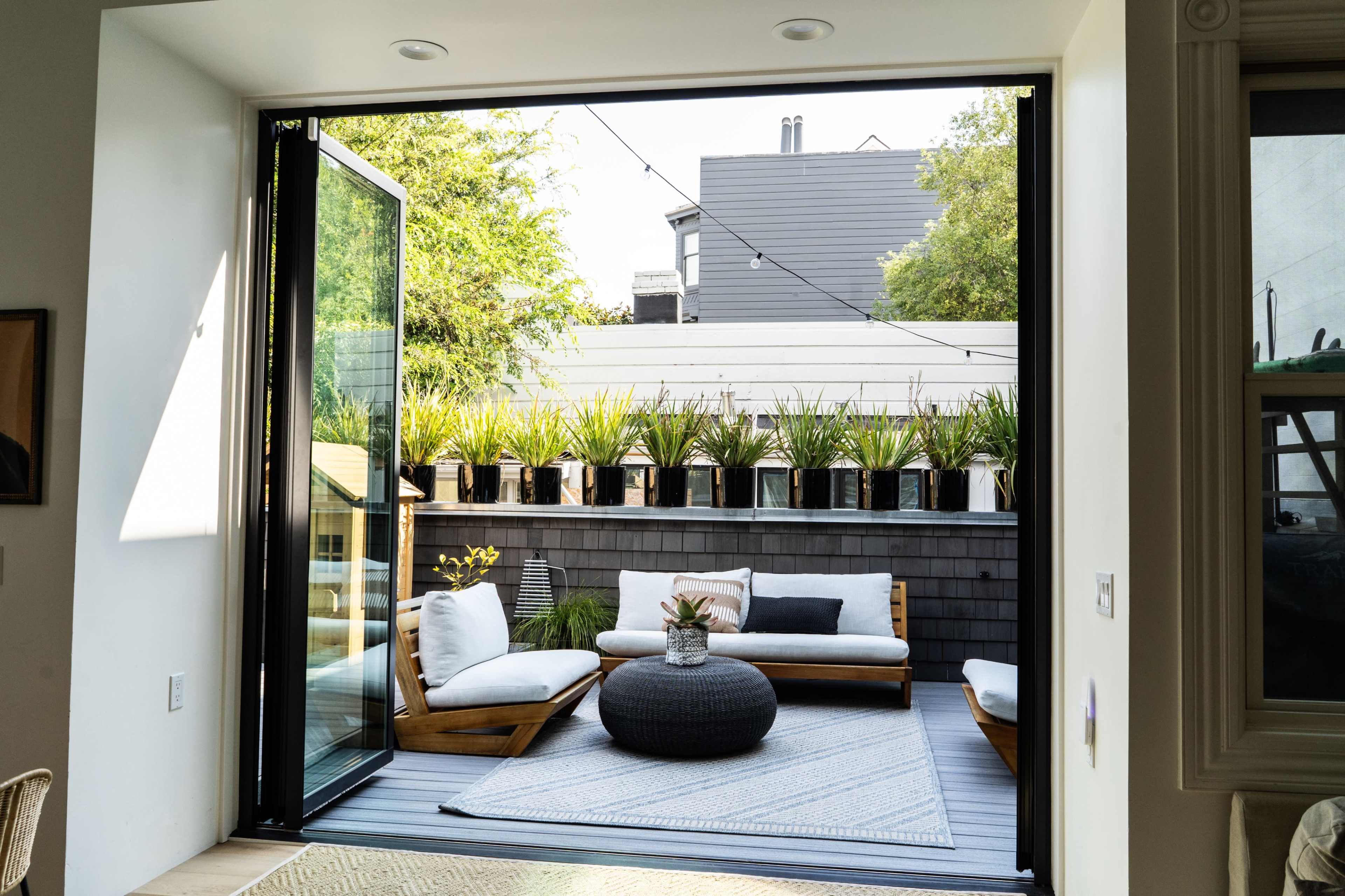A spacious outdoor patio features a sectional sofa, a round ottoman, and a decorative rug, surrounded by potted plants and an open sliding door.