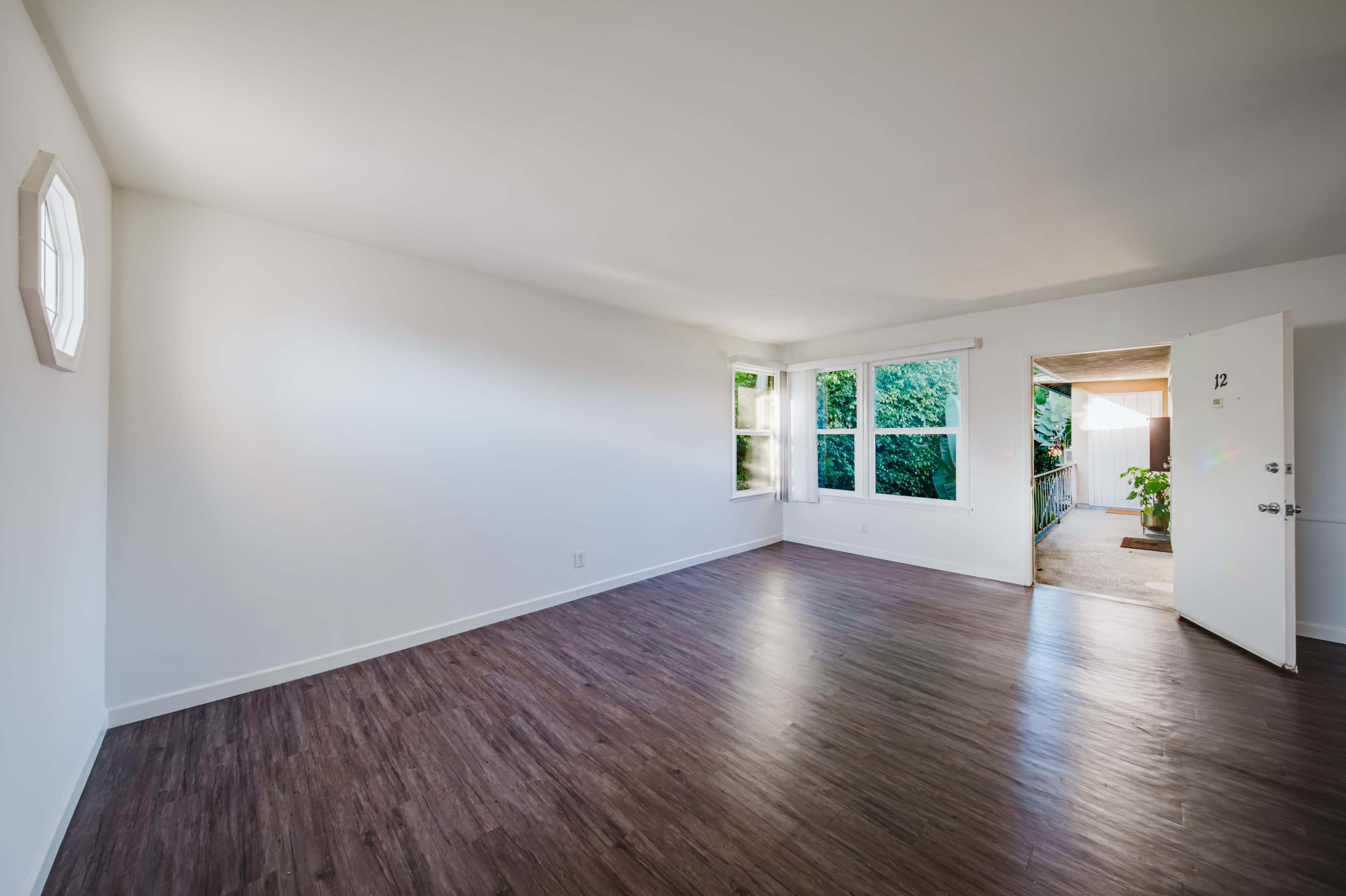 The image shows an empty room with wood-like flooring, a large window with natural light, and a door leading outside.