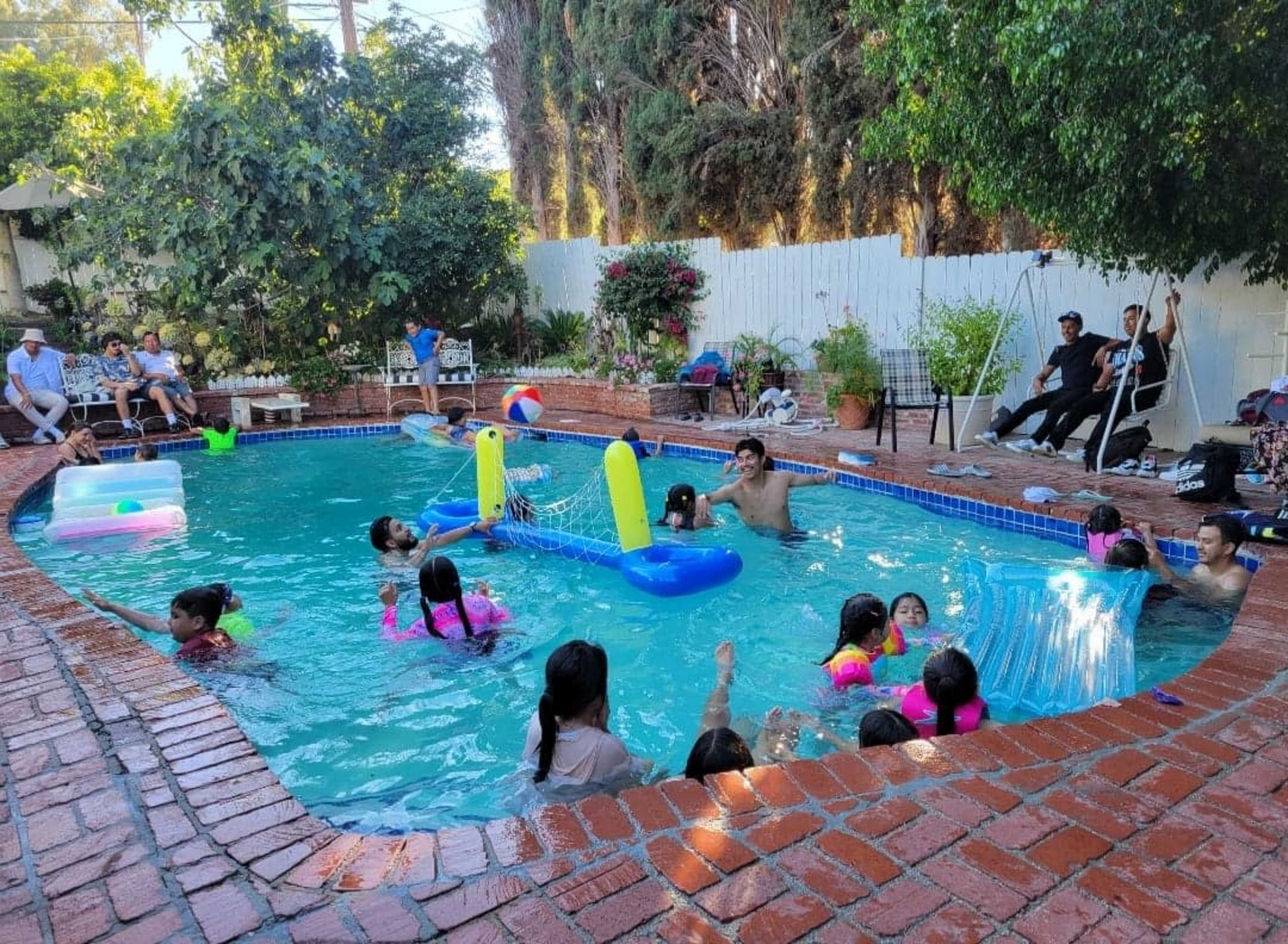 A group of children and adults are gathered around a residential pool filled with inflatable toys, enjoying the warm weather.