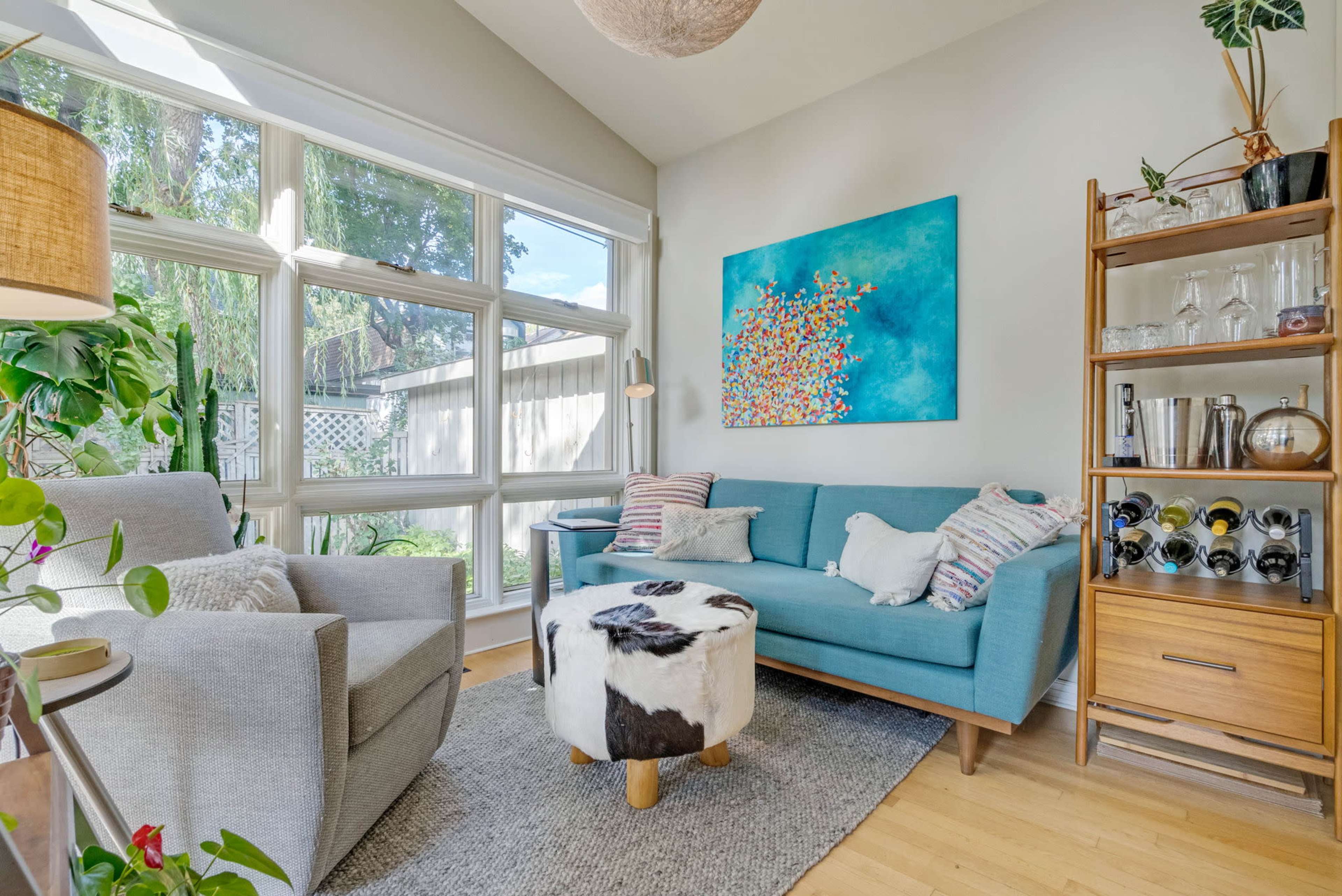A cozy living room features large windows, modern furniture, and a colorful abstract painting on the wall.