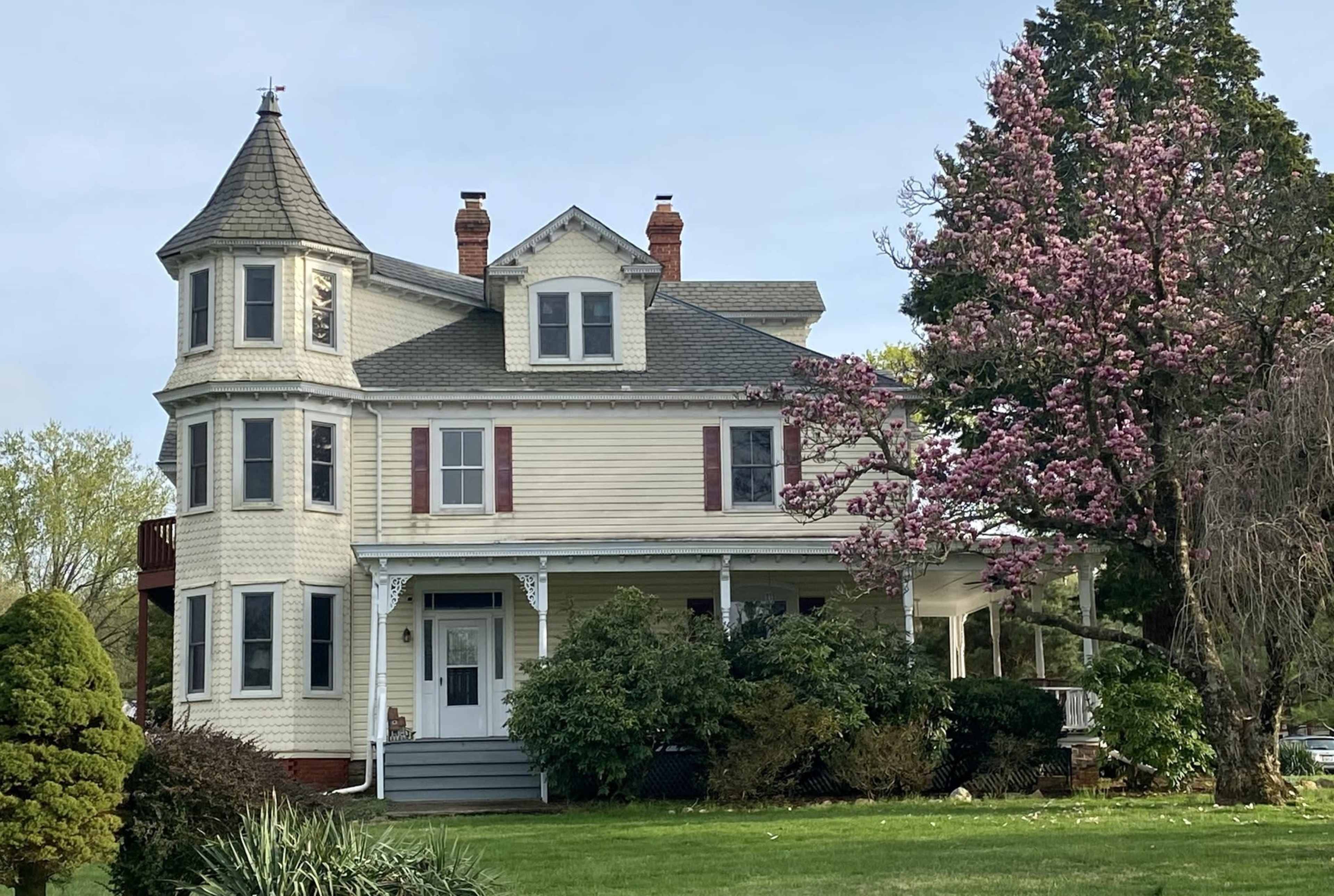 A large, Victorian-style house with a turret and a flowering tree in the front yard.