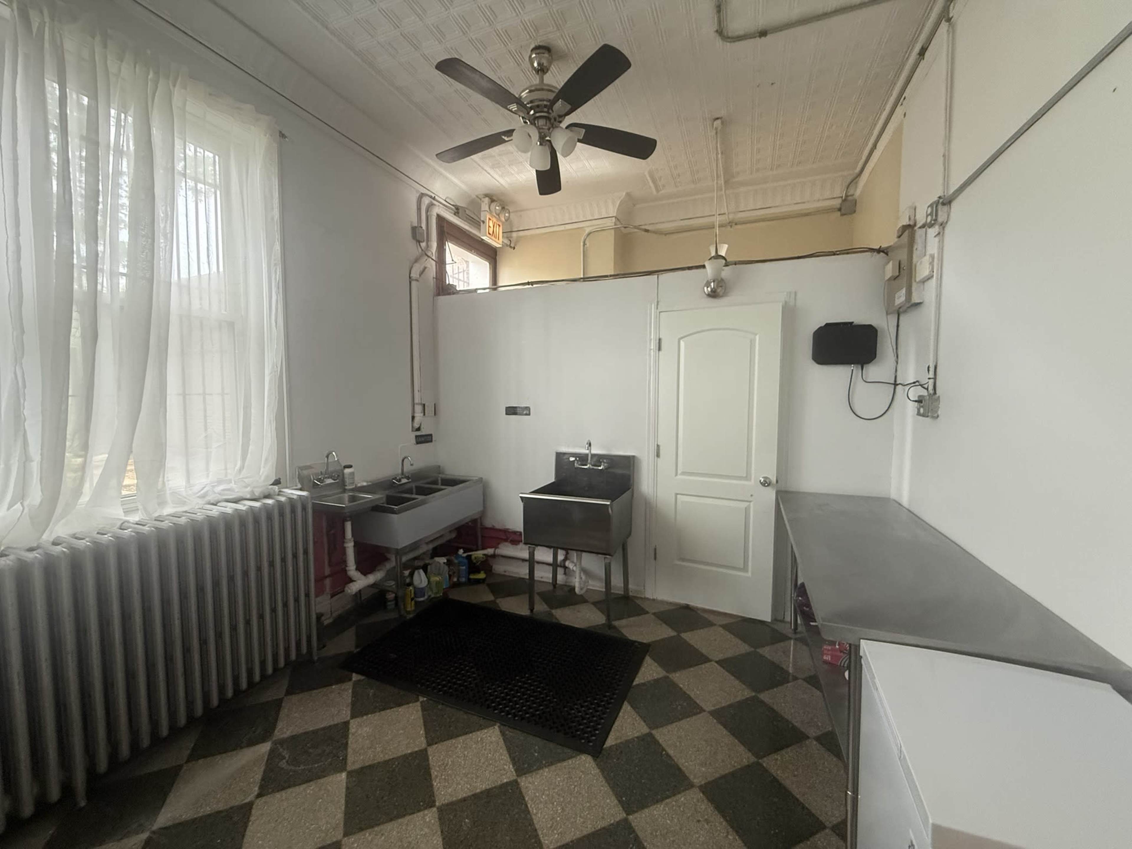 The image shows a kitchen with a ceiling fan, a sink area, a worktable, and a window draped with white curtains.