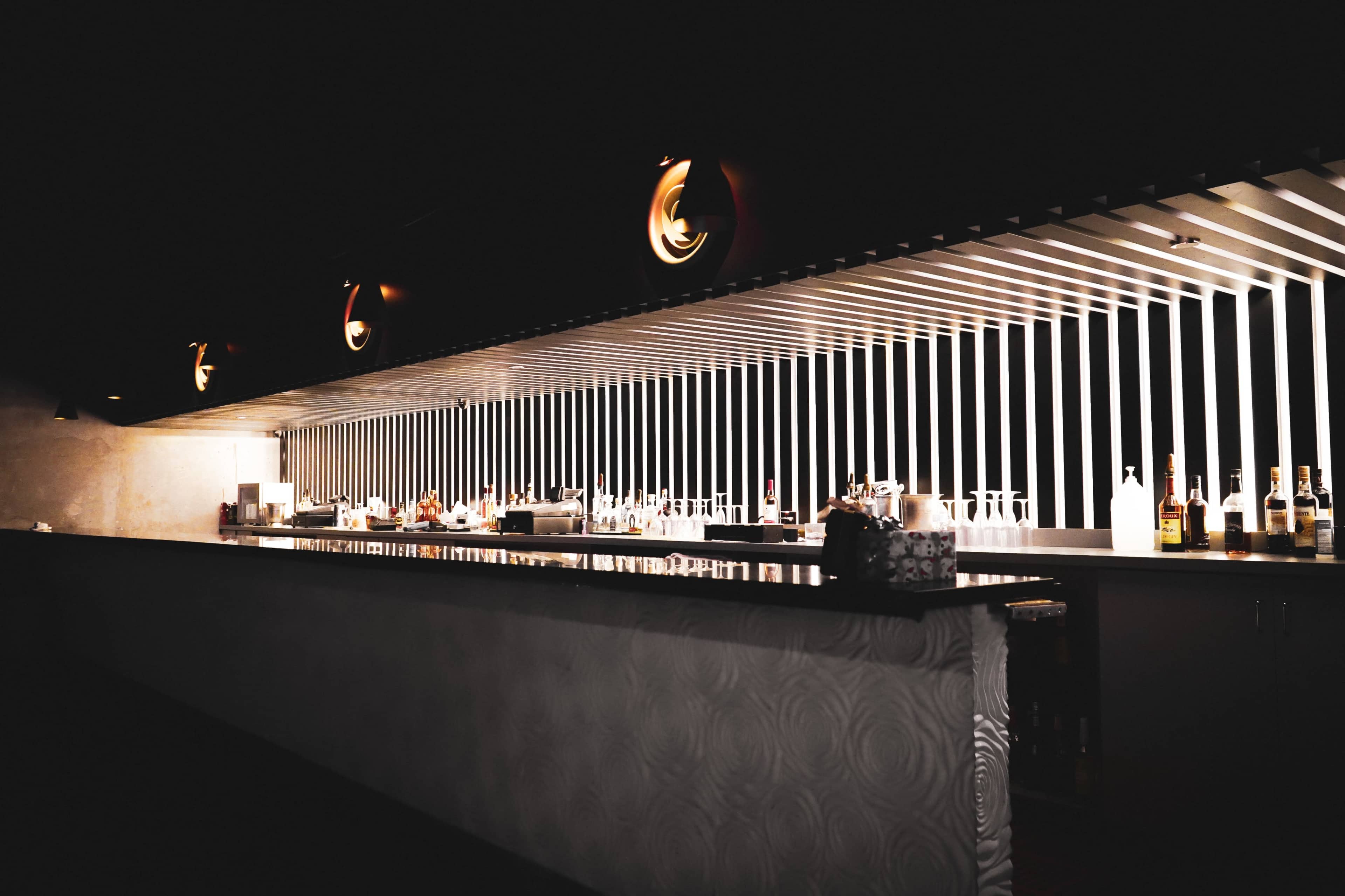 A modern bar with a sleek counter and illuminated shelves displaying various bottles.