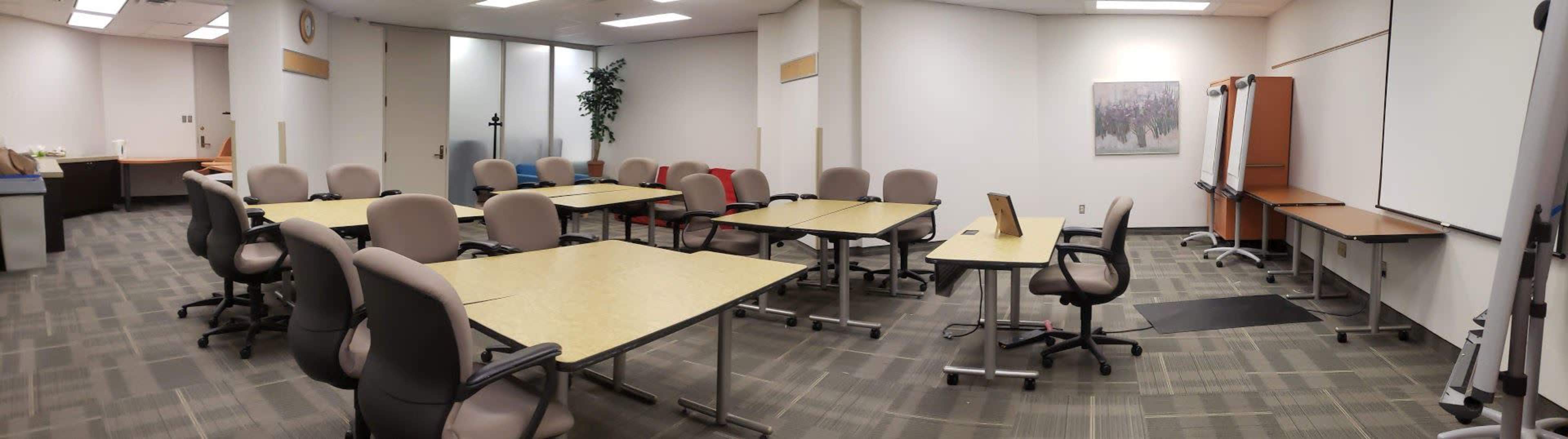 A conference room with several tables and chairs arranged for a meeting, along with a whiteboard and wall-mounted screen.