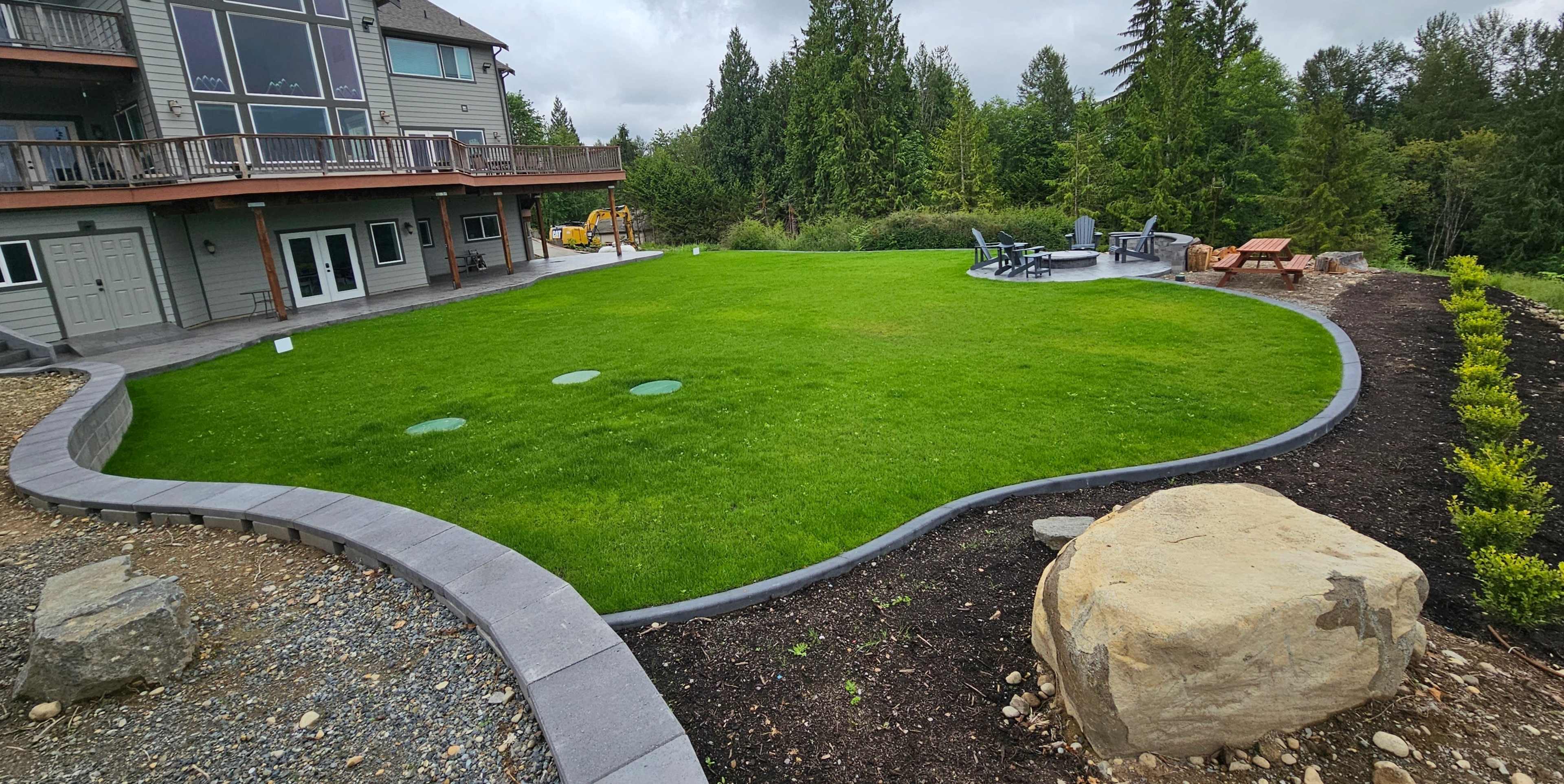 The image shows a landscaped backyard featuring a green lawn with a curved stone border, a seating area with outdoor furniture, and a view of trees in the background.