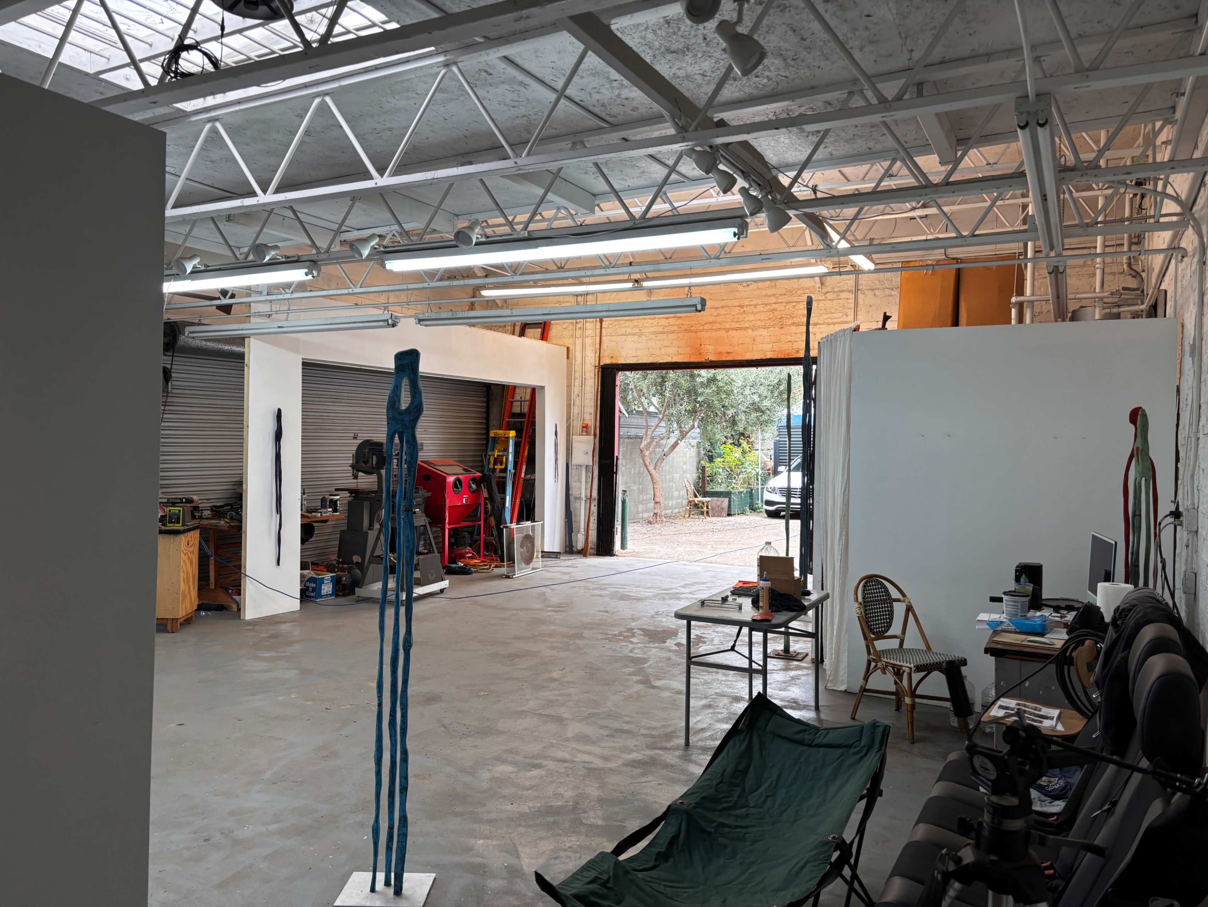 The image shows an open workshop with large white partitions, artwork displayed, and a view of an outdoor area through a wide doorway.