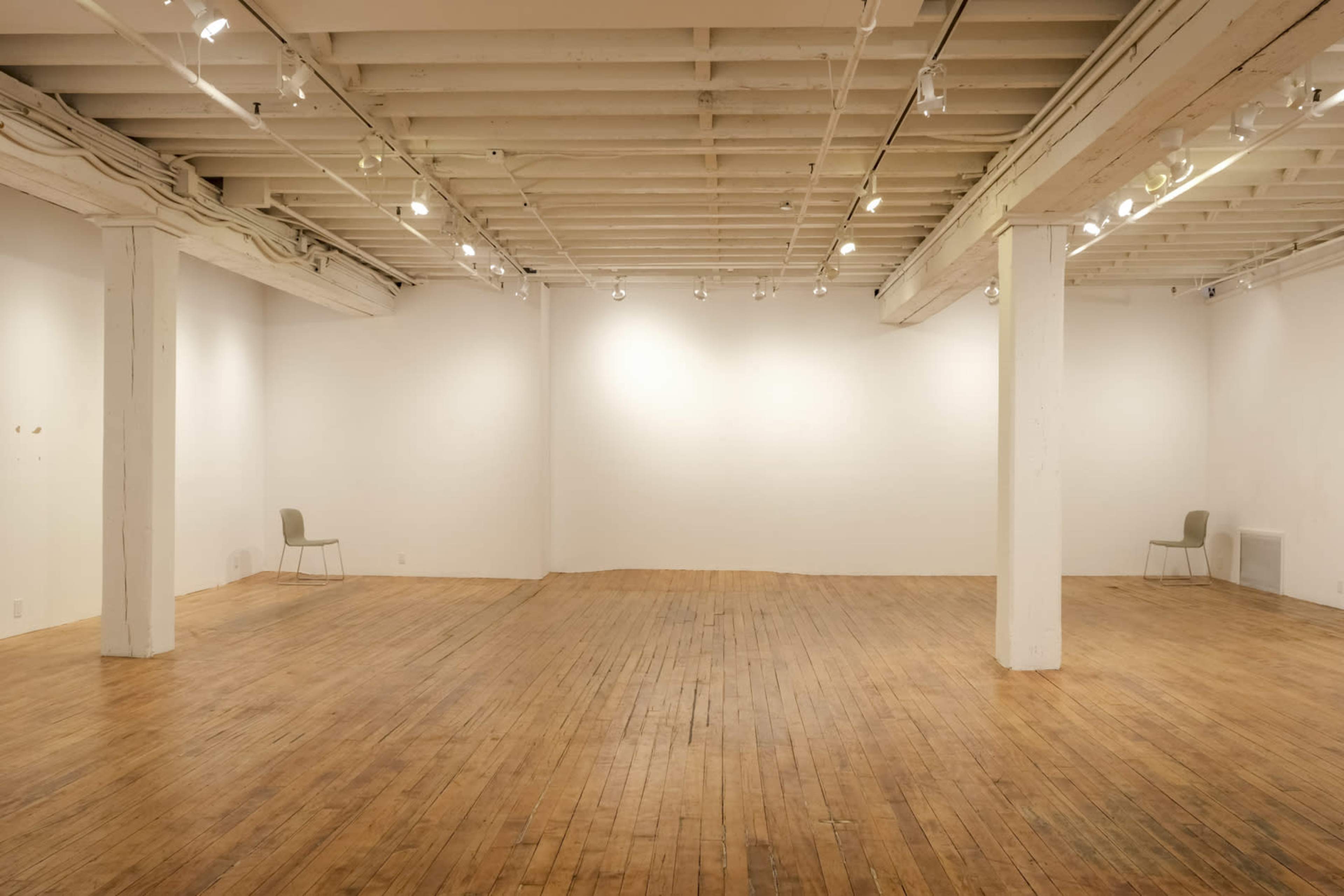 An empty art gallery features wooden flooring, white walls, and two gray chairs placed on opposite sides of the room.