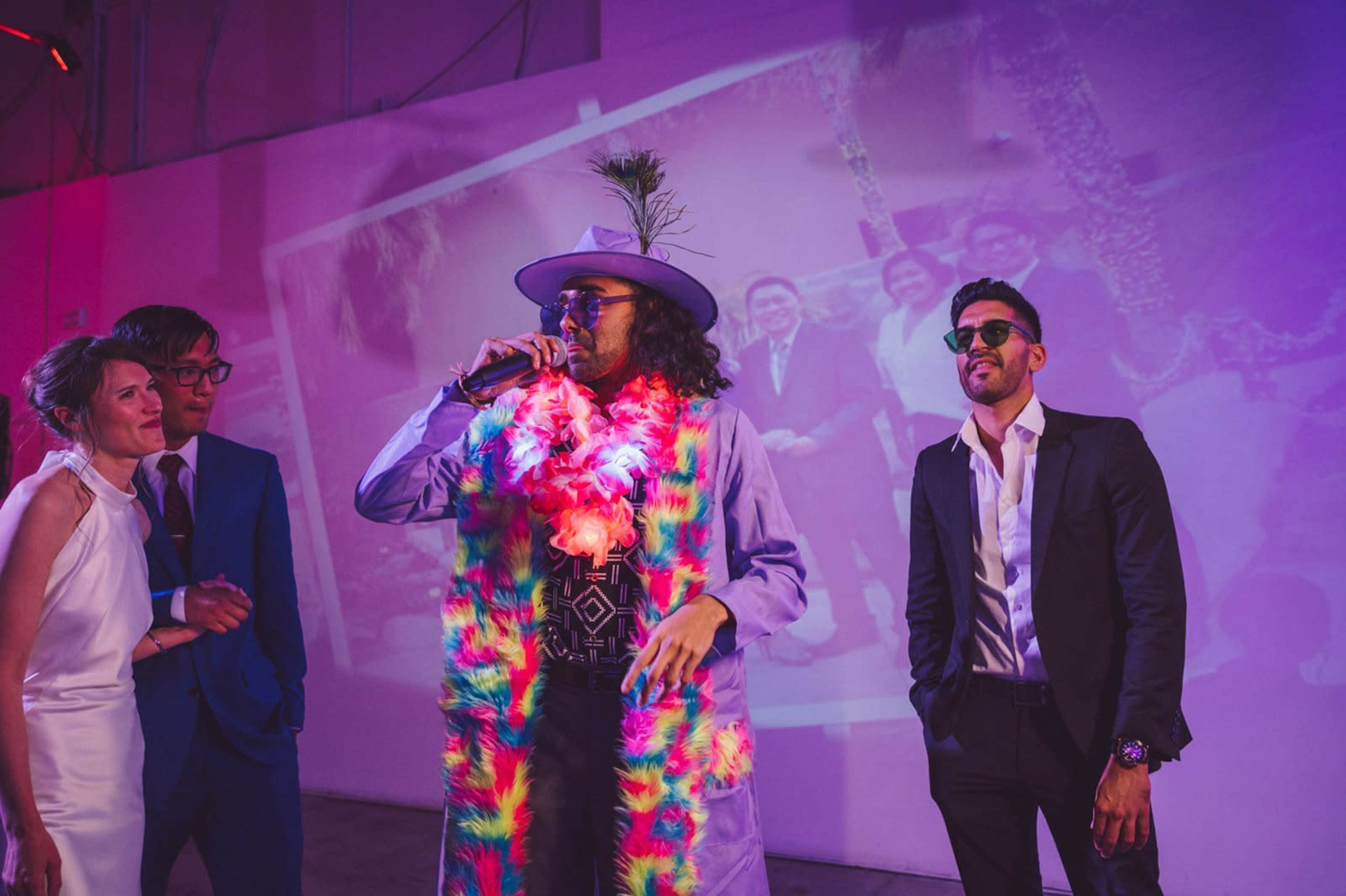 A person in a colorful outfit holds a microphone while standing in front of a group of people at a lively event.