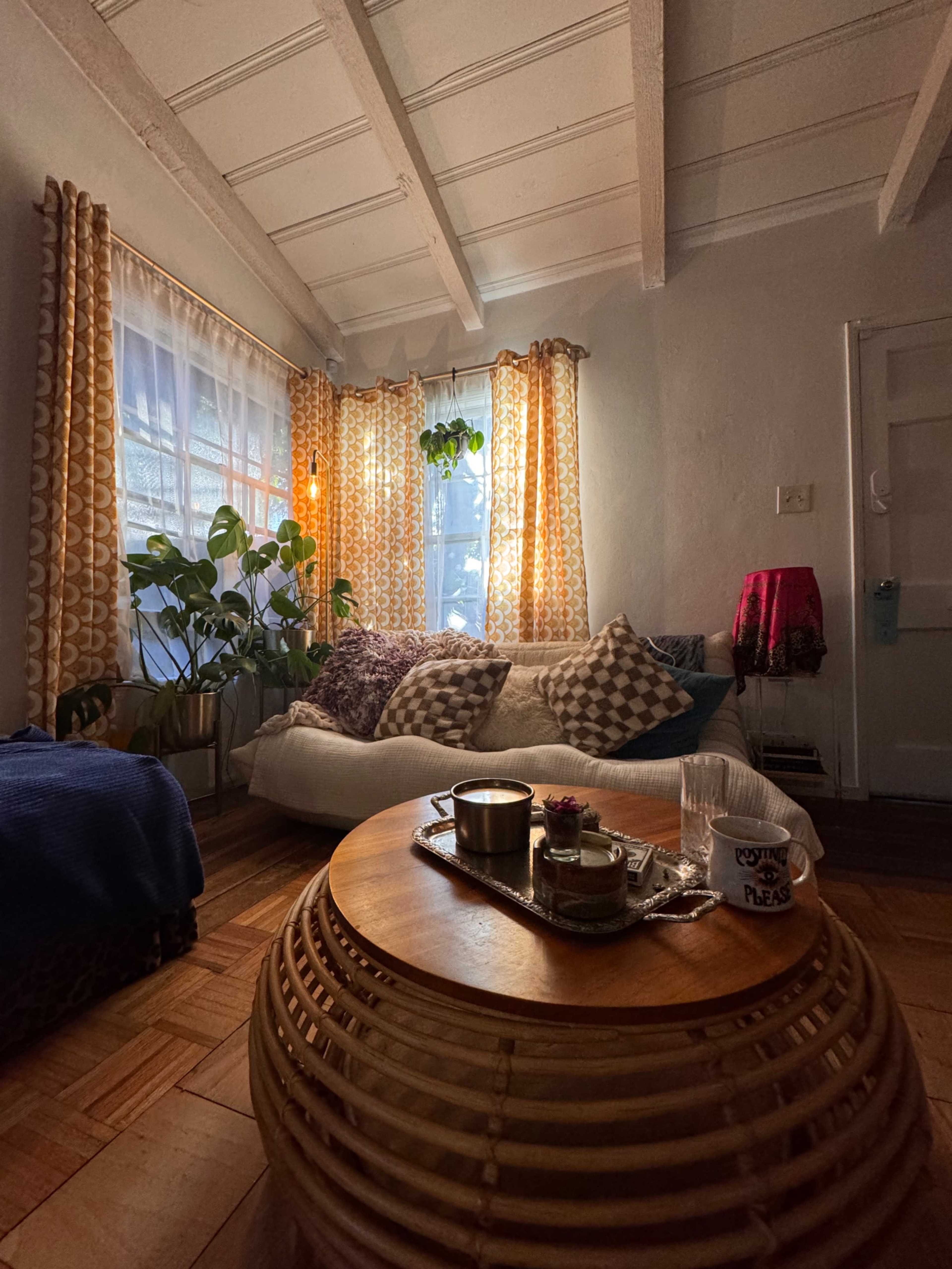 A cozy living room with a sofa adorned with patterned pillows, a round wooden coffee table, and plants near a window with sheer curtains.
