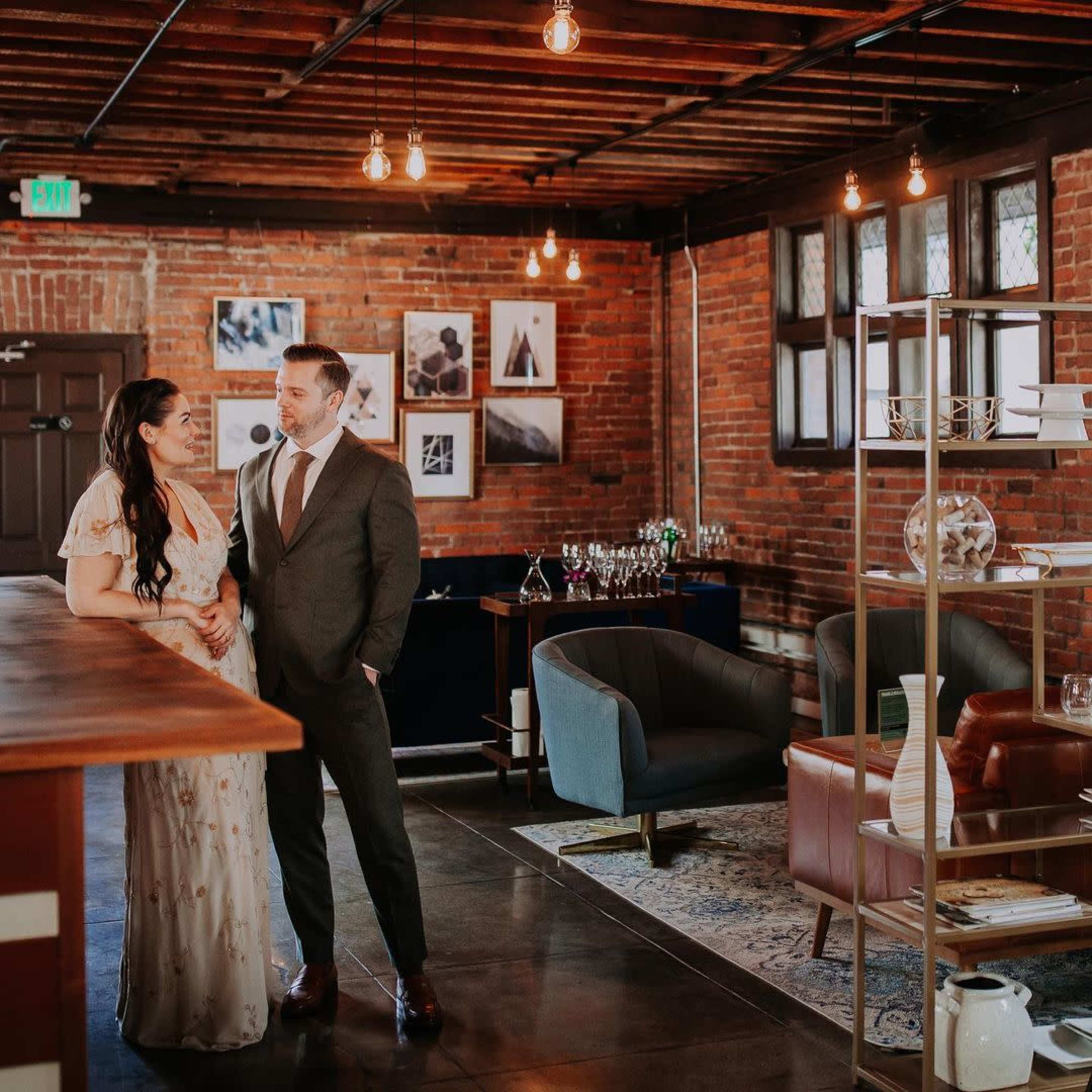 A couple stands together in a stylish venue with exposed brick walls, modern furnishings, and soft overhead lighting.