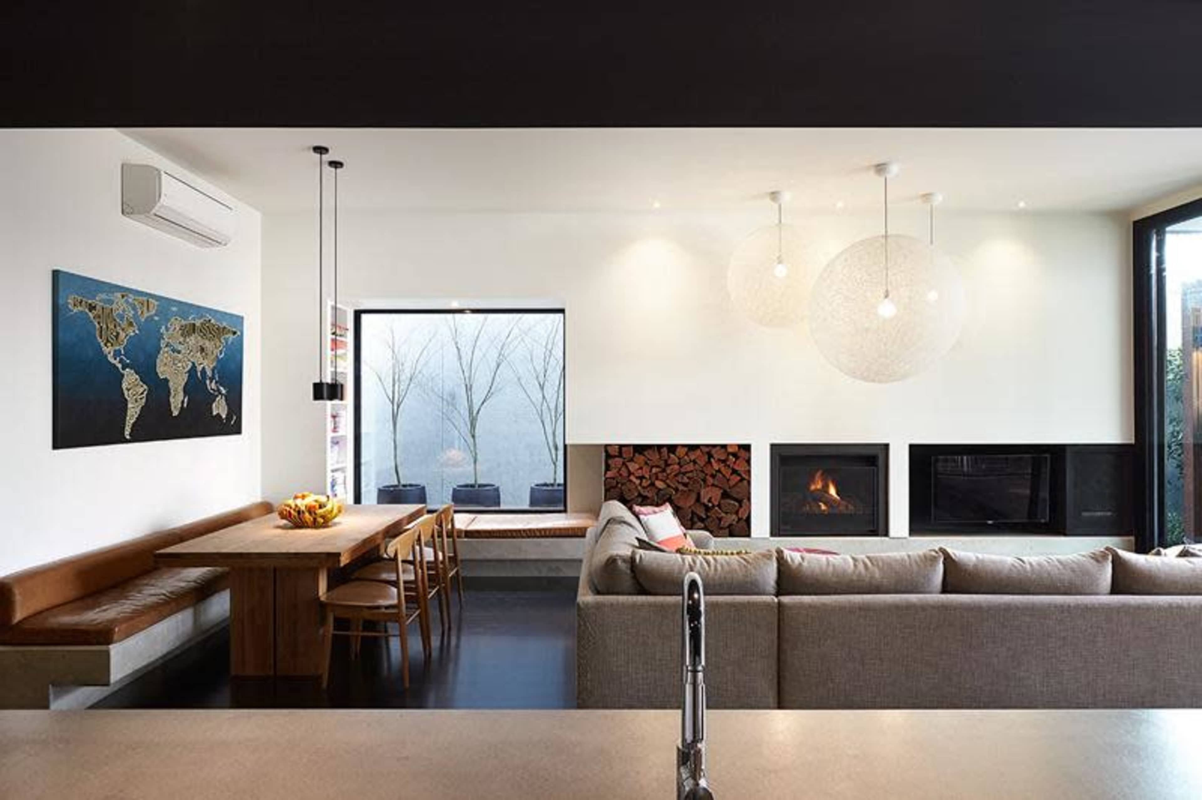 A modern living space featuring a dining area with a wooden table and chairs, a cozy sofa, a fireplace with stacked wood, and a large window providing natural light.