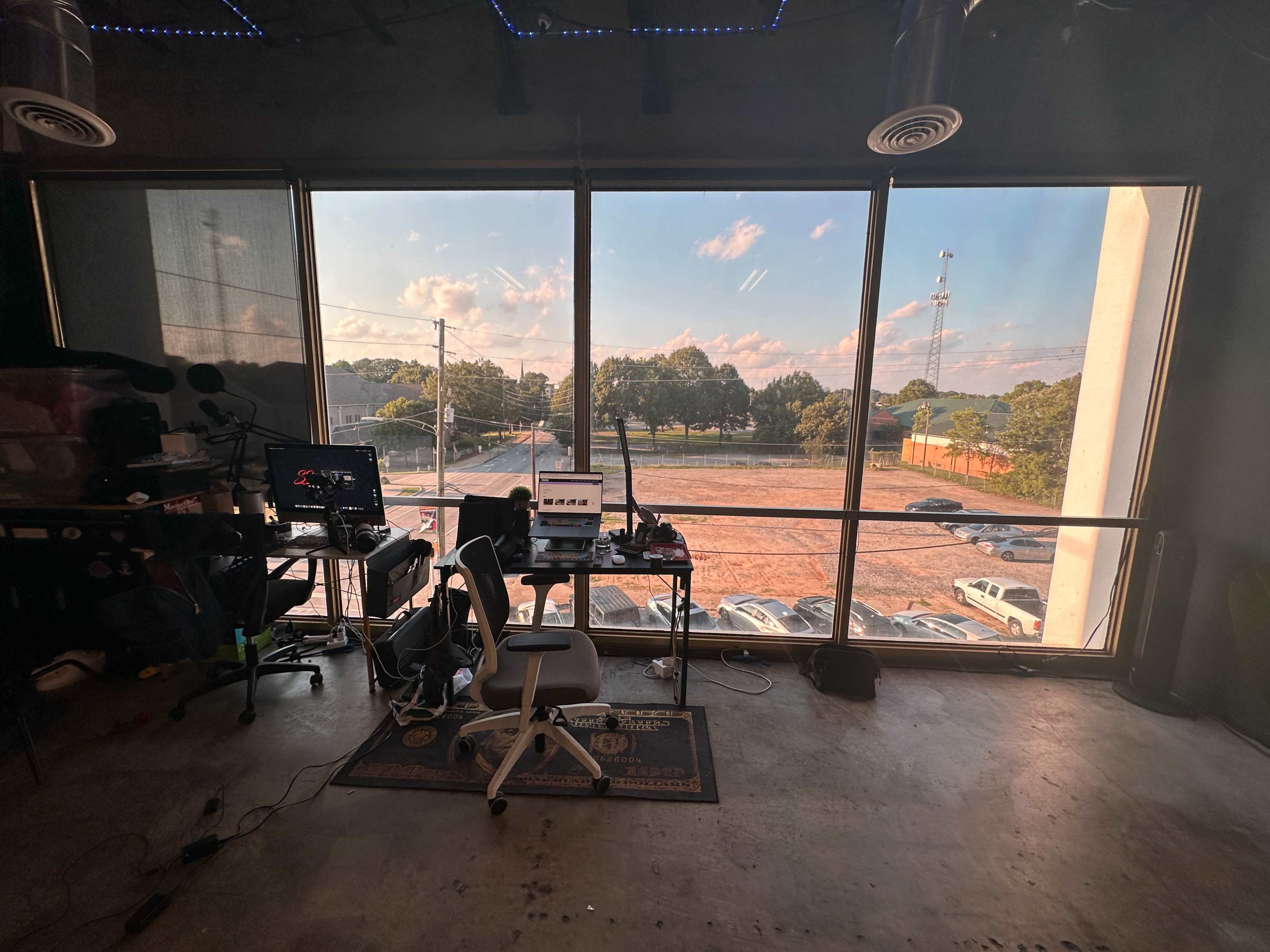 An office space with a desk, computer setup, and a large window overlooking a parking area and trees outside.
