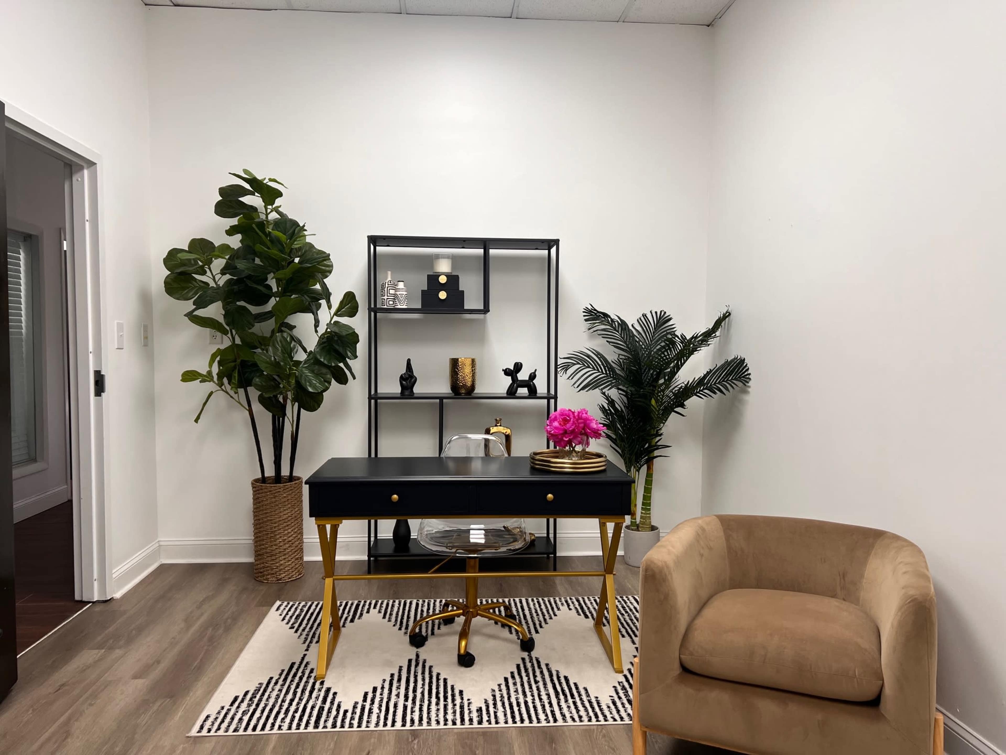 The image shows a modern office space featuring a black desk with gold accents, a beige armchair, and decorative plants alongside a bookshelf with various decorative items.