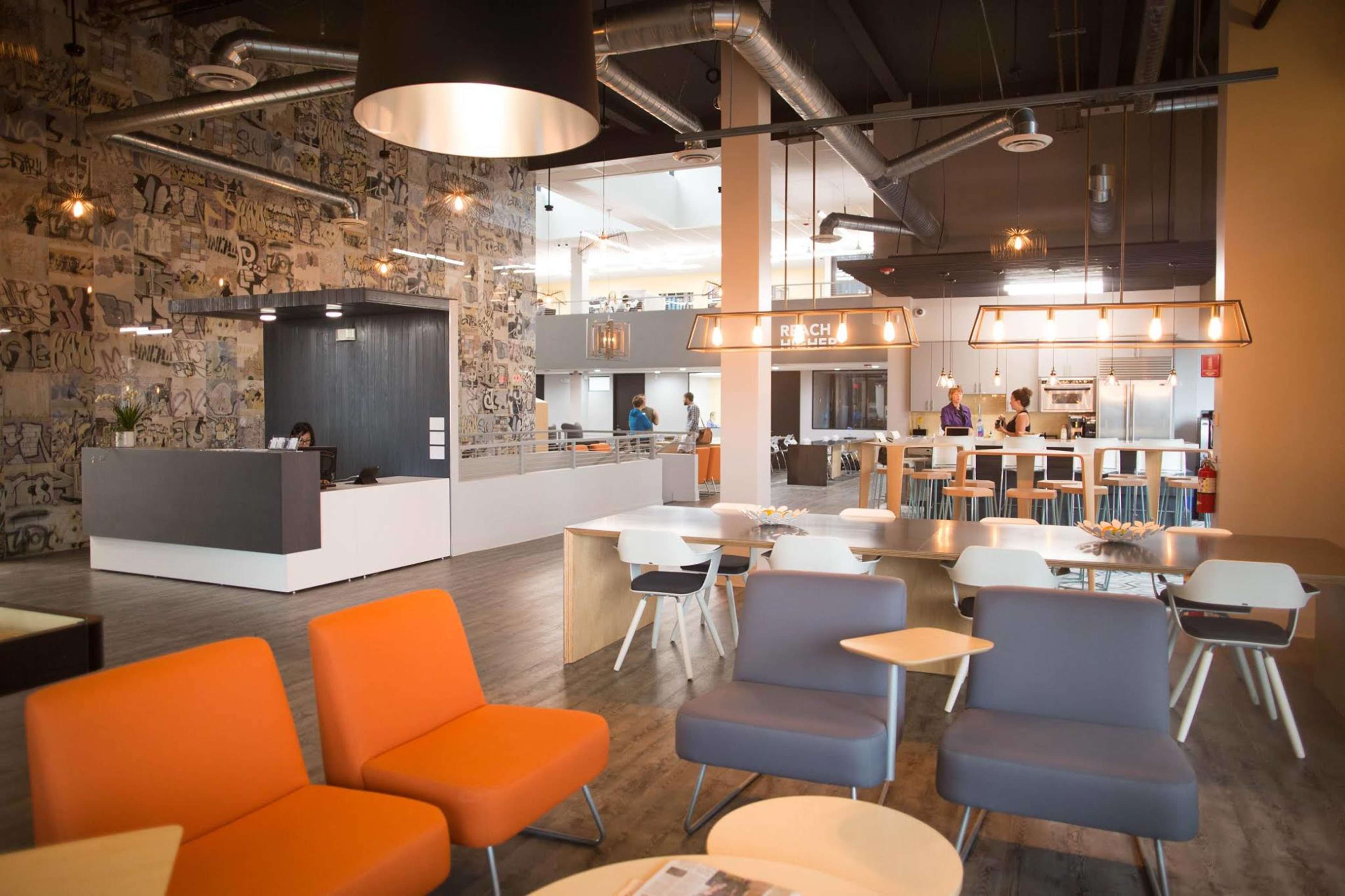 A modern office interior with a reception area, seating arrangements in orange and gray, a kitchen area, and a mural on the wall.