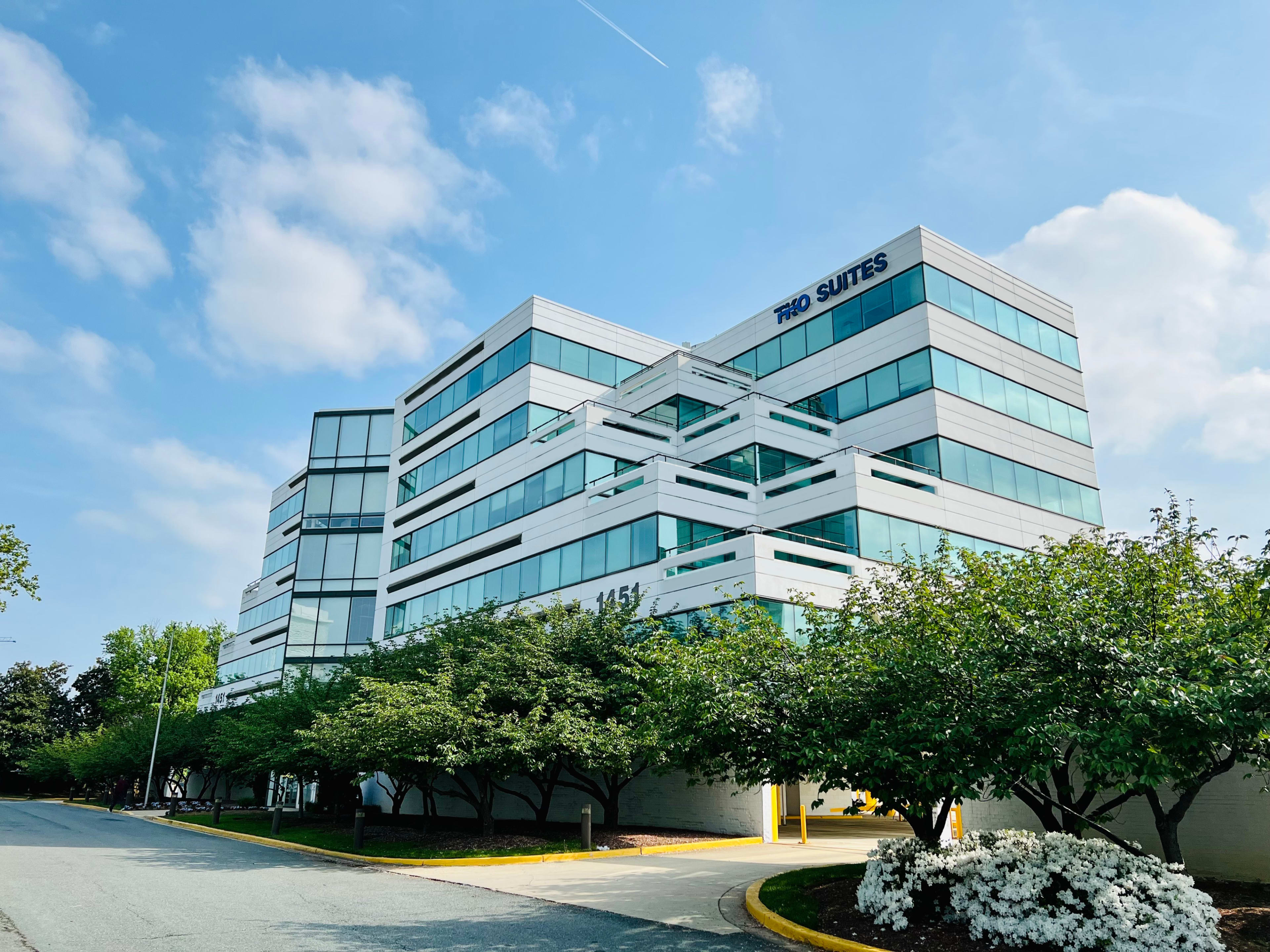 A multi-story office building with large glass windows and a white exterior is surrounded by green landscaping.