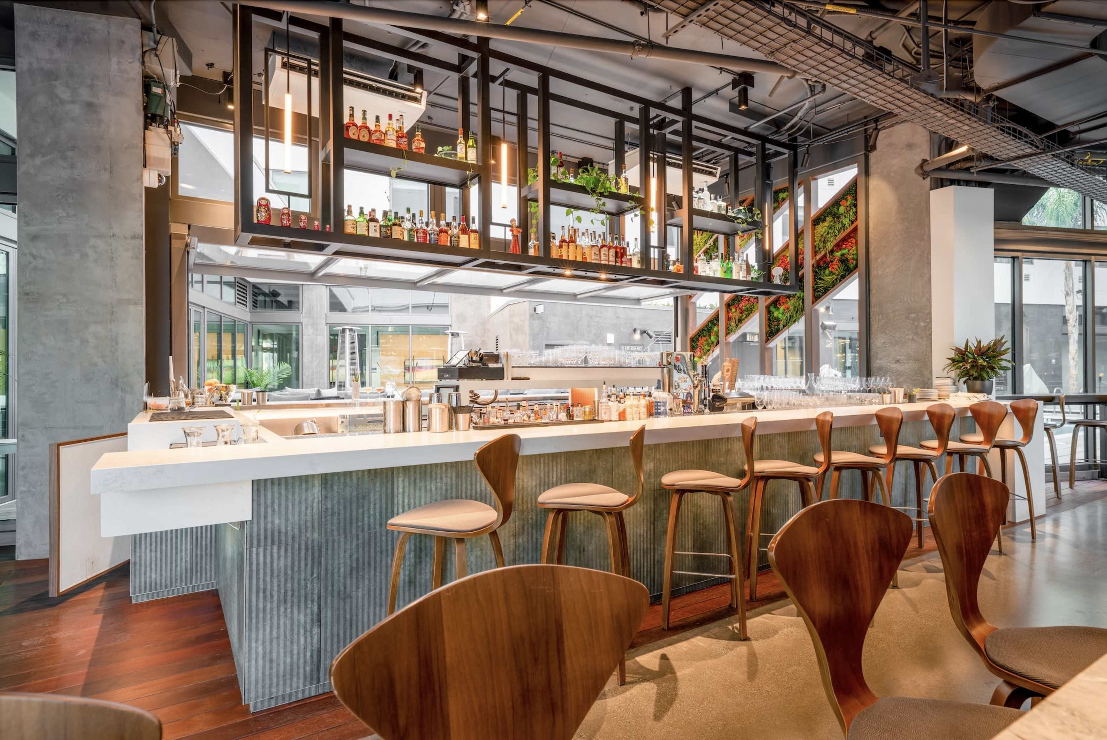 The image shows a modern bar area with a clean white countertop, wooden stools, and shelves displaying various bottles and greenery.