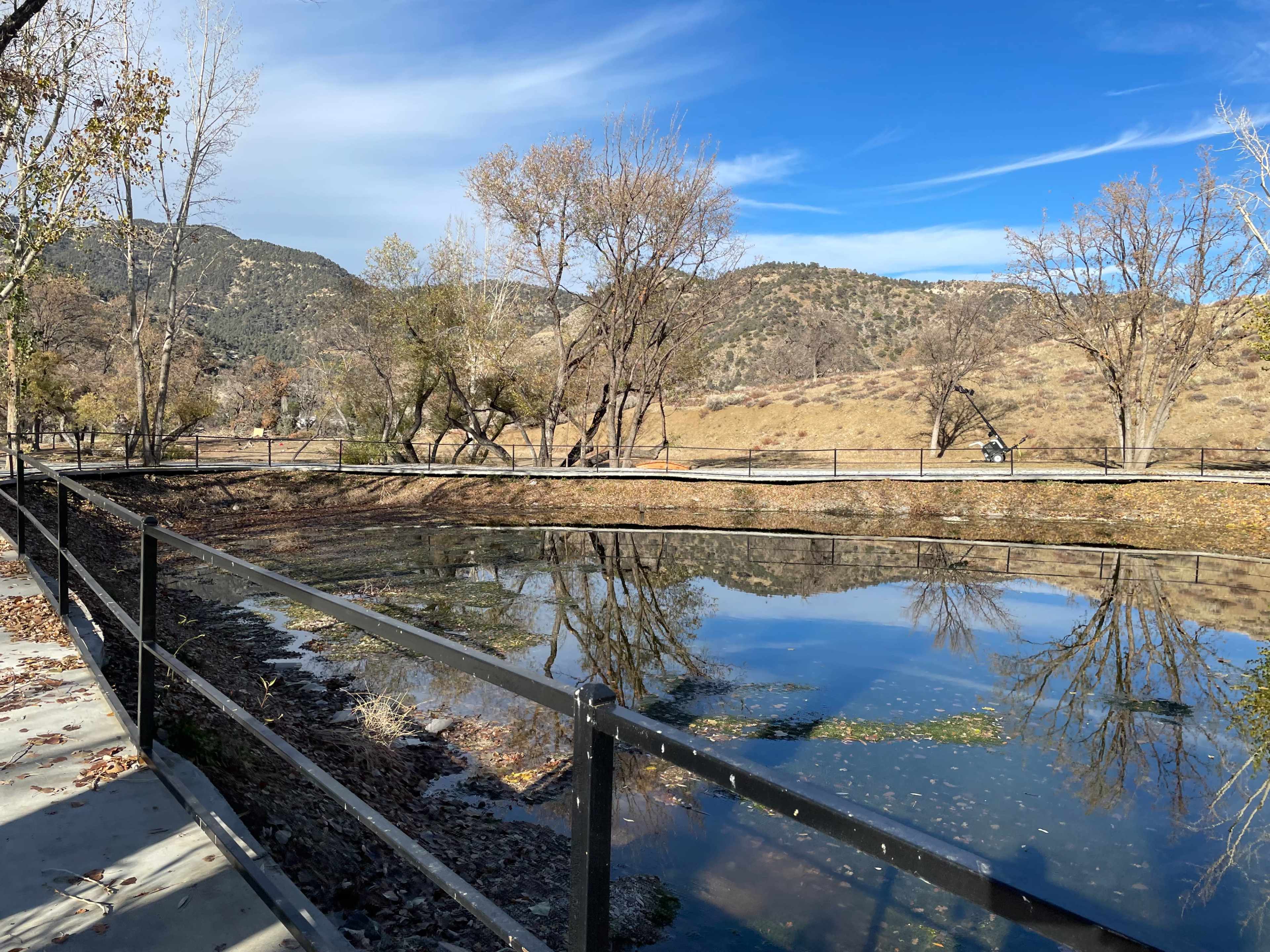 A calm pond reflects the surrounding trees and mountains under a clear blue sky.