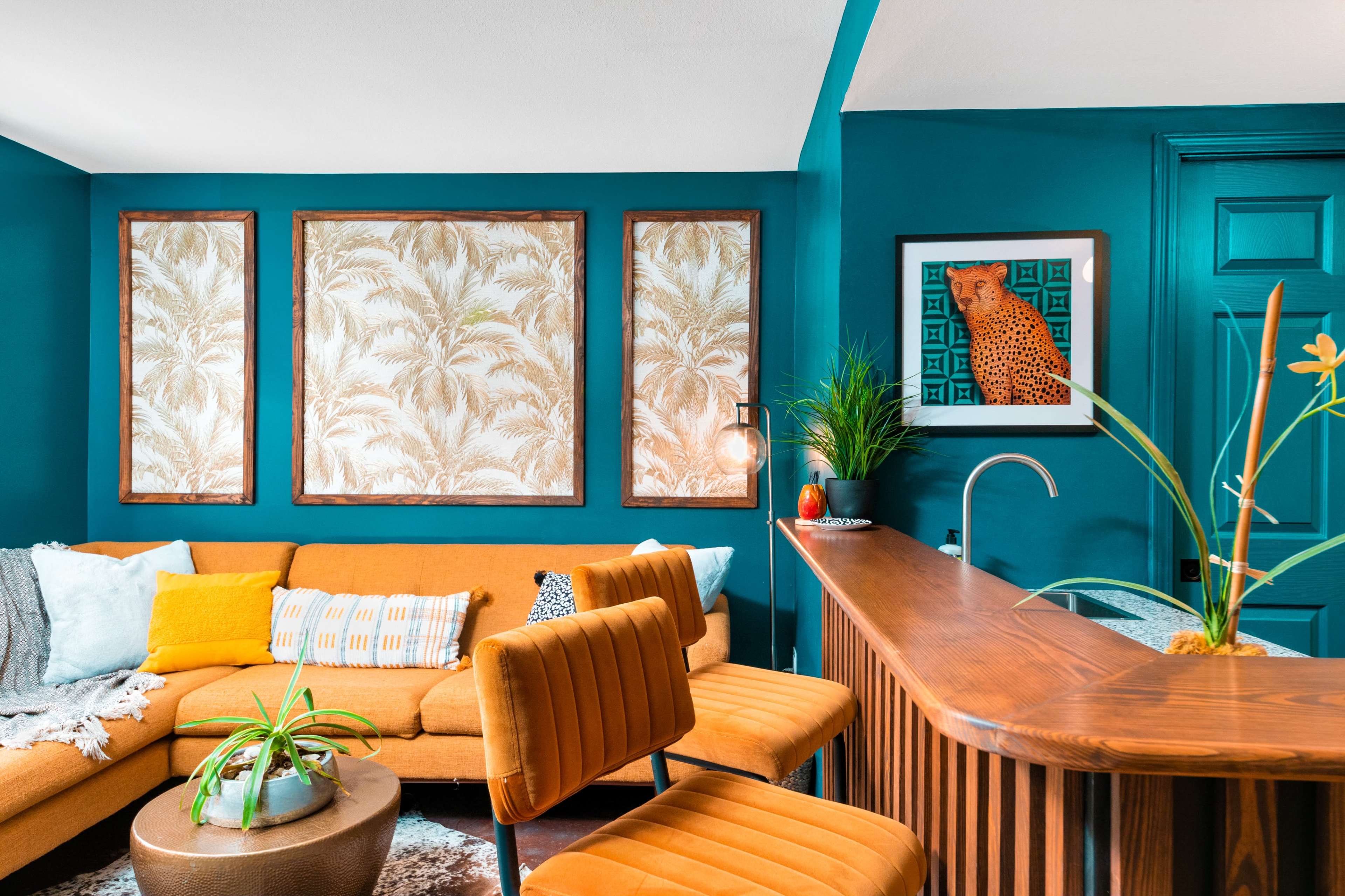 A bright living room featuring a mustard-colored sectional sofa, framed botanical prints on the turquoise walls, and a sleek bar counter with modern seating.