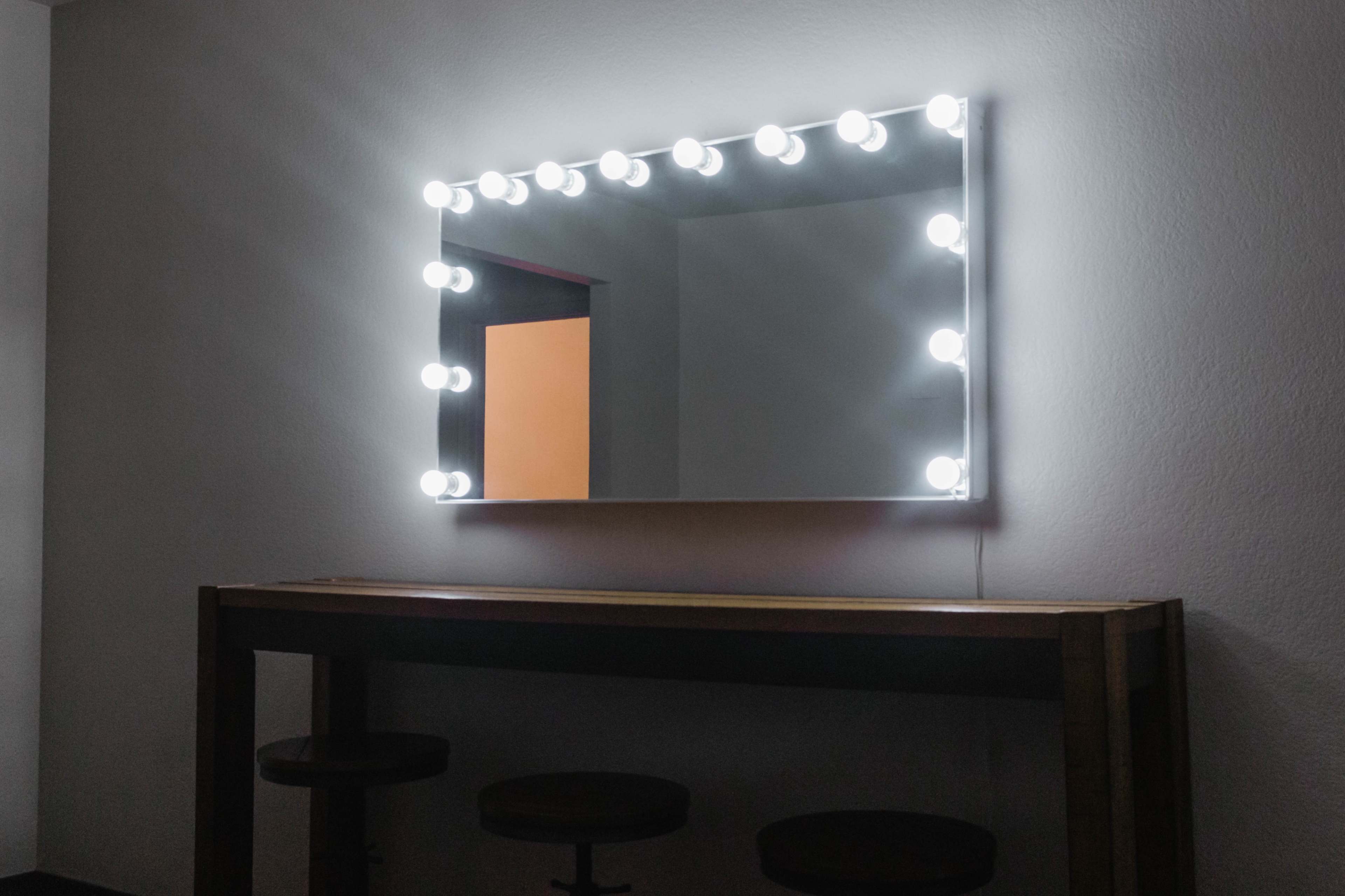 A brightly lit mirror with circular lights surrounds it, mounted on a wall above a wooden table with three stools.