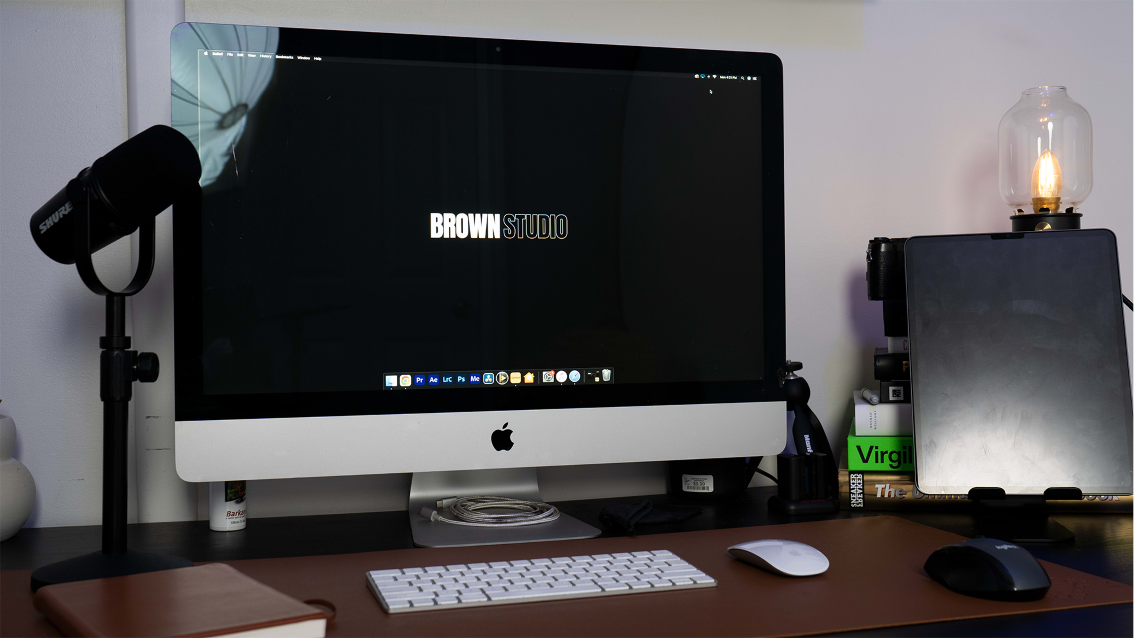 A modern desk setup features an iMac displaying a logo, a microphone, a notebook, and a lamp.