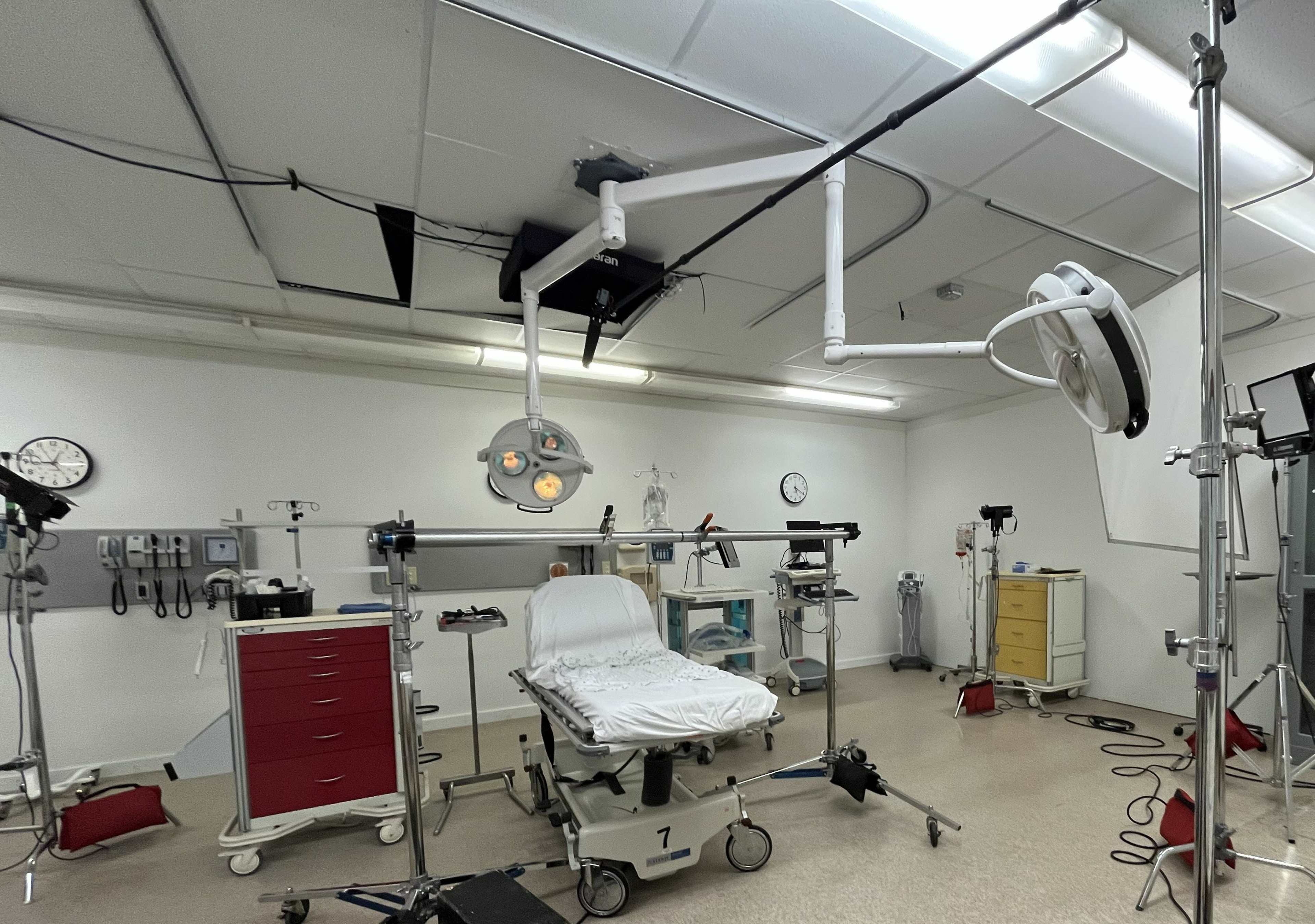 The image shows an operating room with a surgical table, overhead lights, medical equipment, and a variety of instruments arranged around the space.