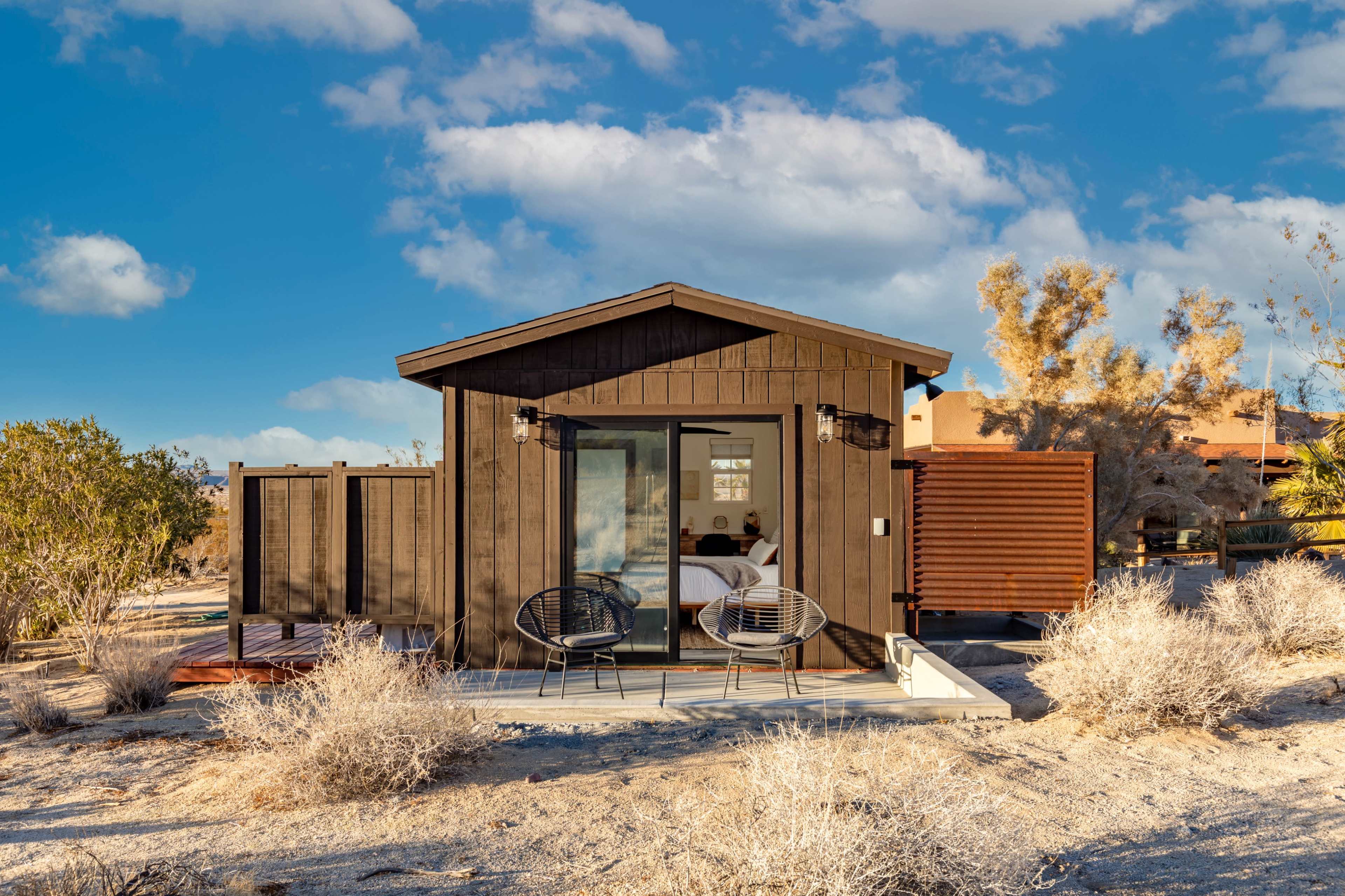 A modern black cabin with a deck and two chairs is set in a desolate landscape with sparse vegetation and clear blue skies.