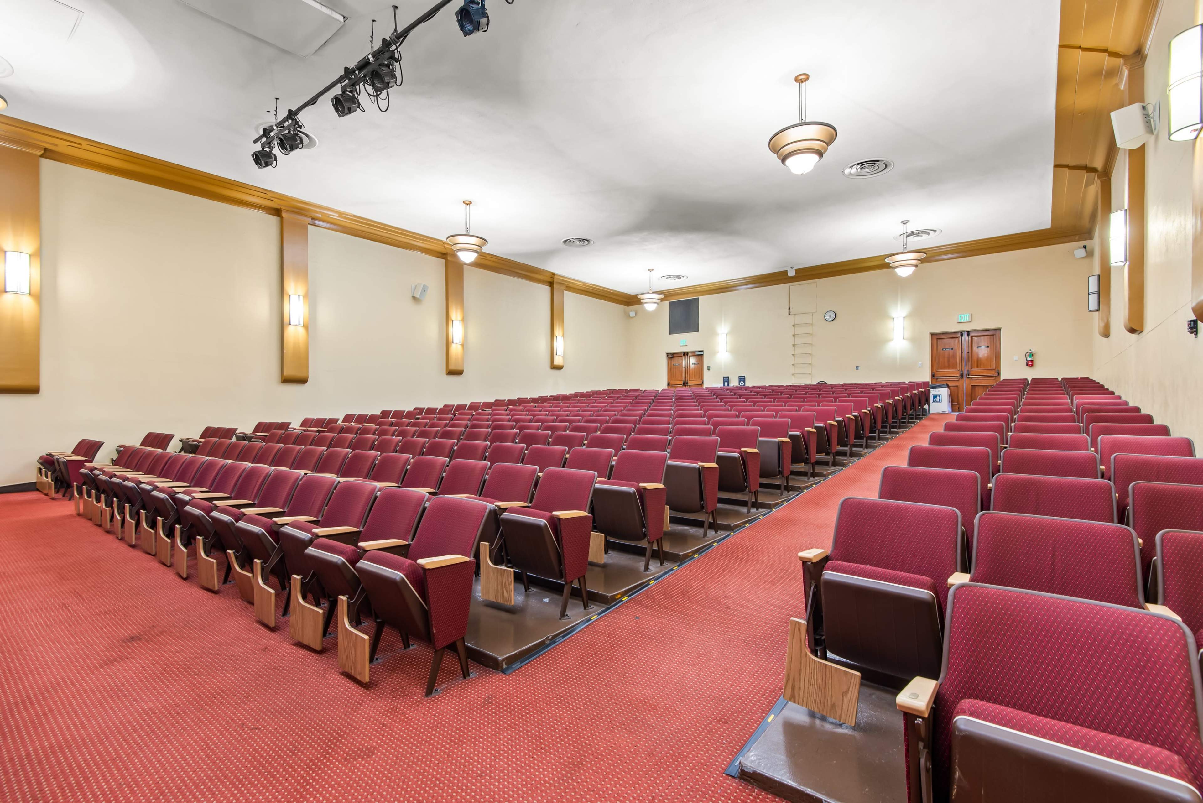 The image depicts a spacious auditorium with rows of maroon seating, a red carpeted floor, and stage lighting fixtures overhead.