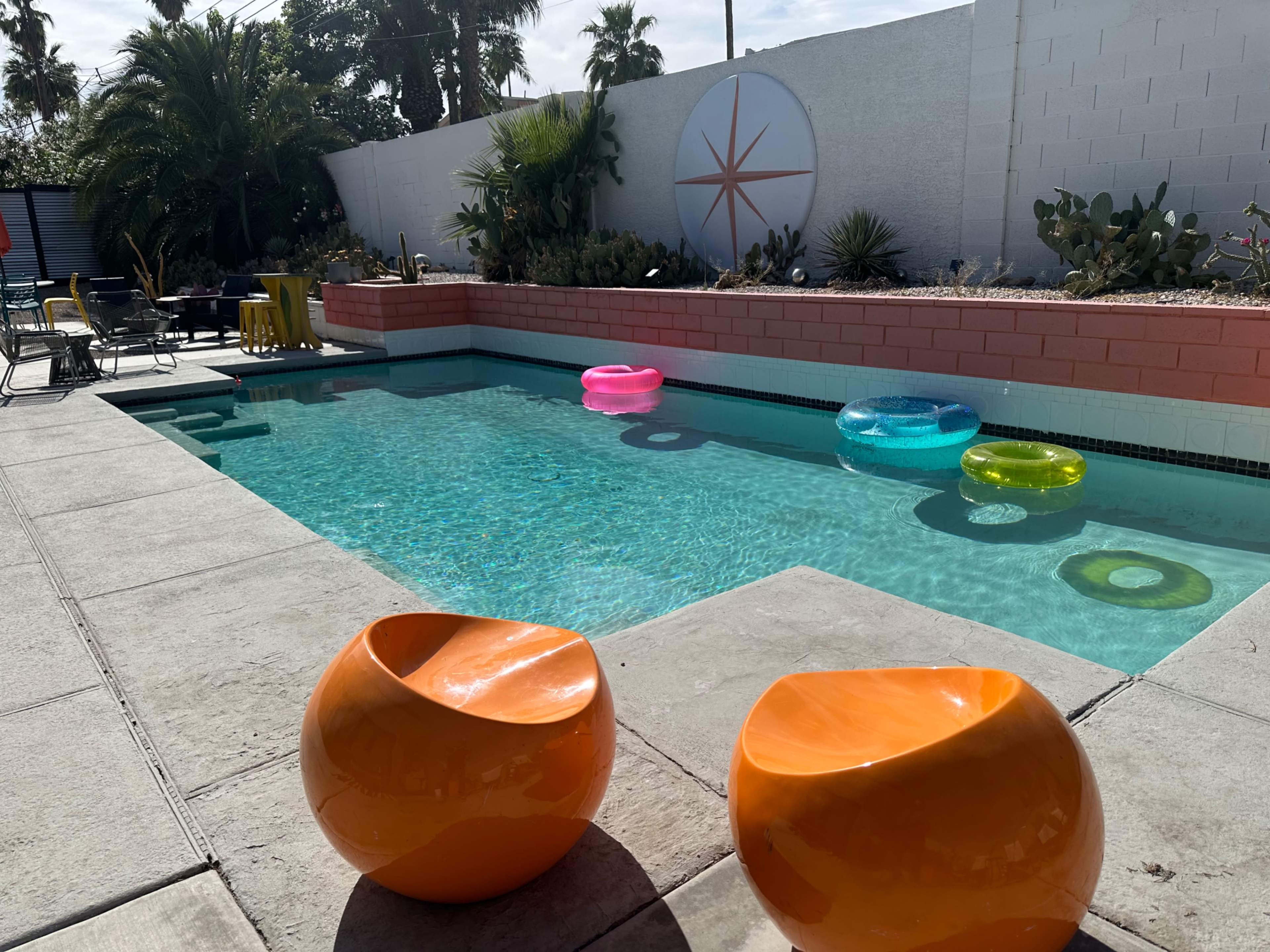 The image shows a swimming pool surrounded by concrete, featuring colorful inflatable rings and orange chairs nearby.