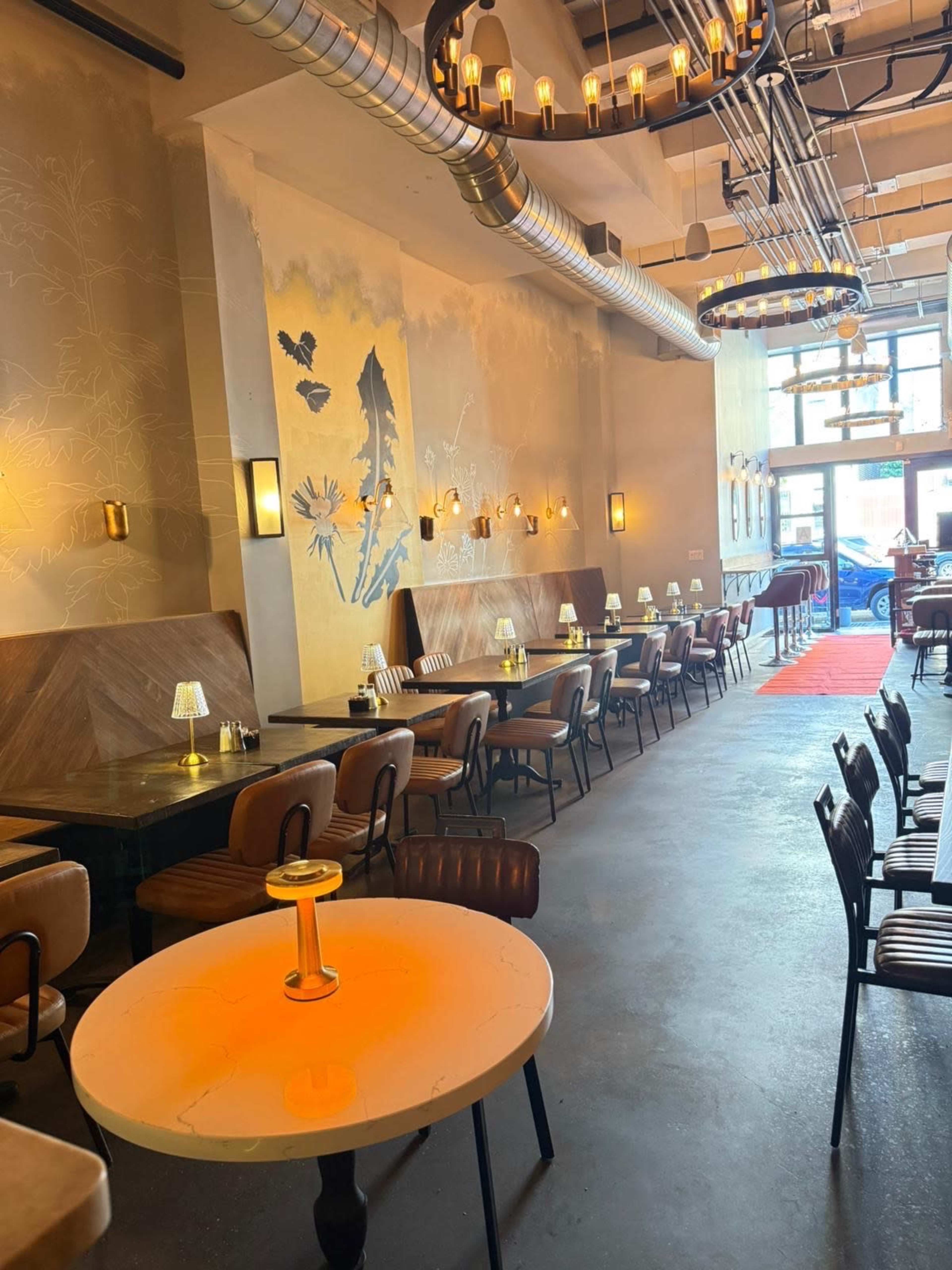 The image shows a stylish restaurant interior with wooden seating, small tables, and decorative lighting, featuring a textured wall mural and large windows that let in natural light.