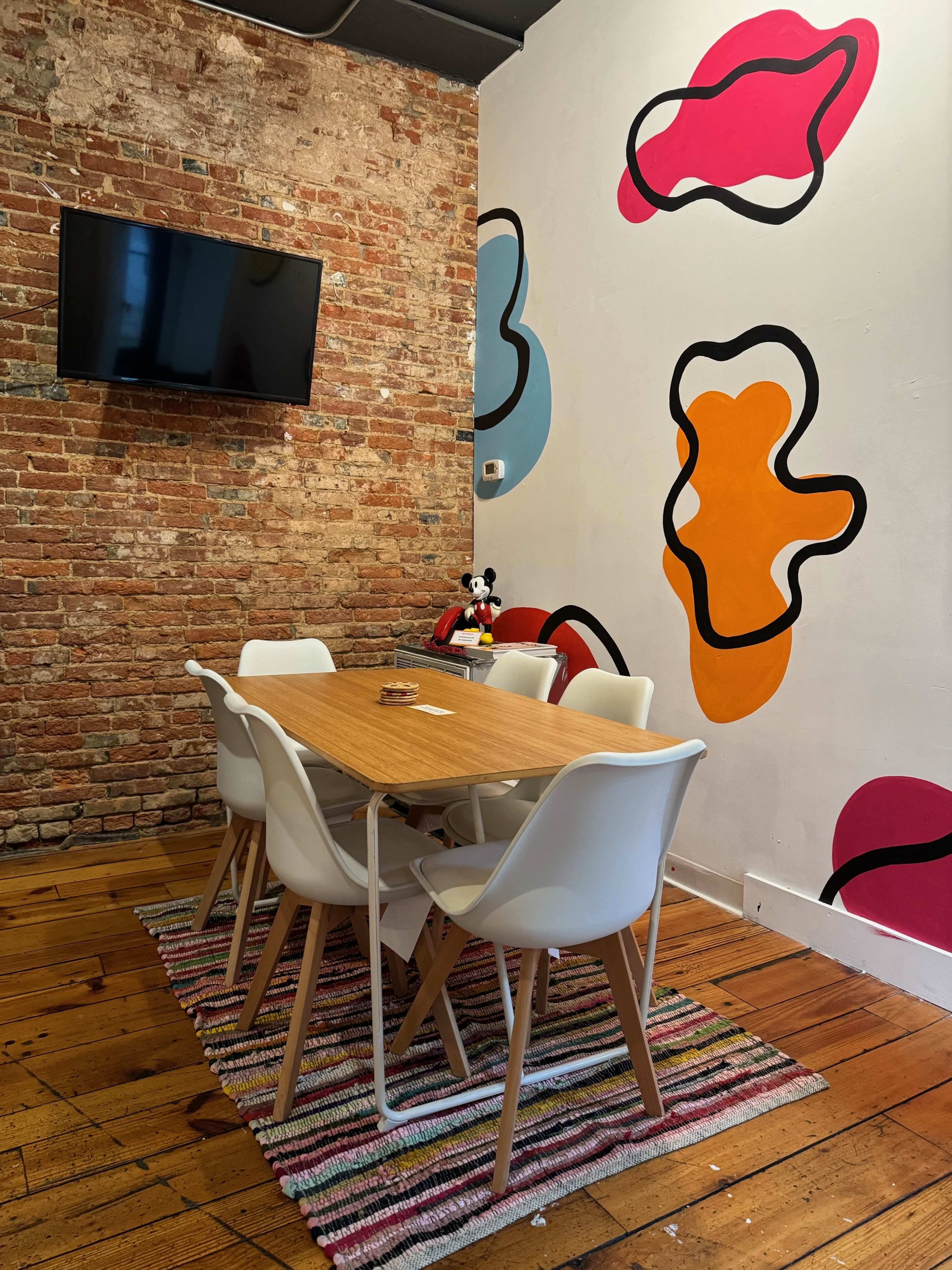 A modern meeting room with a wooden table surrounded by white chairs, a brick wall, and colorful abstract shapes painted on the wall.