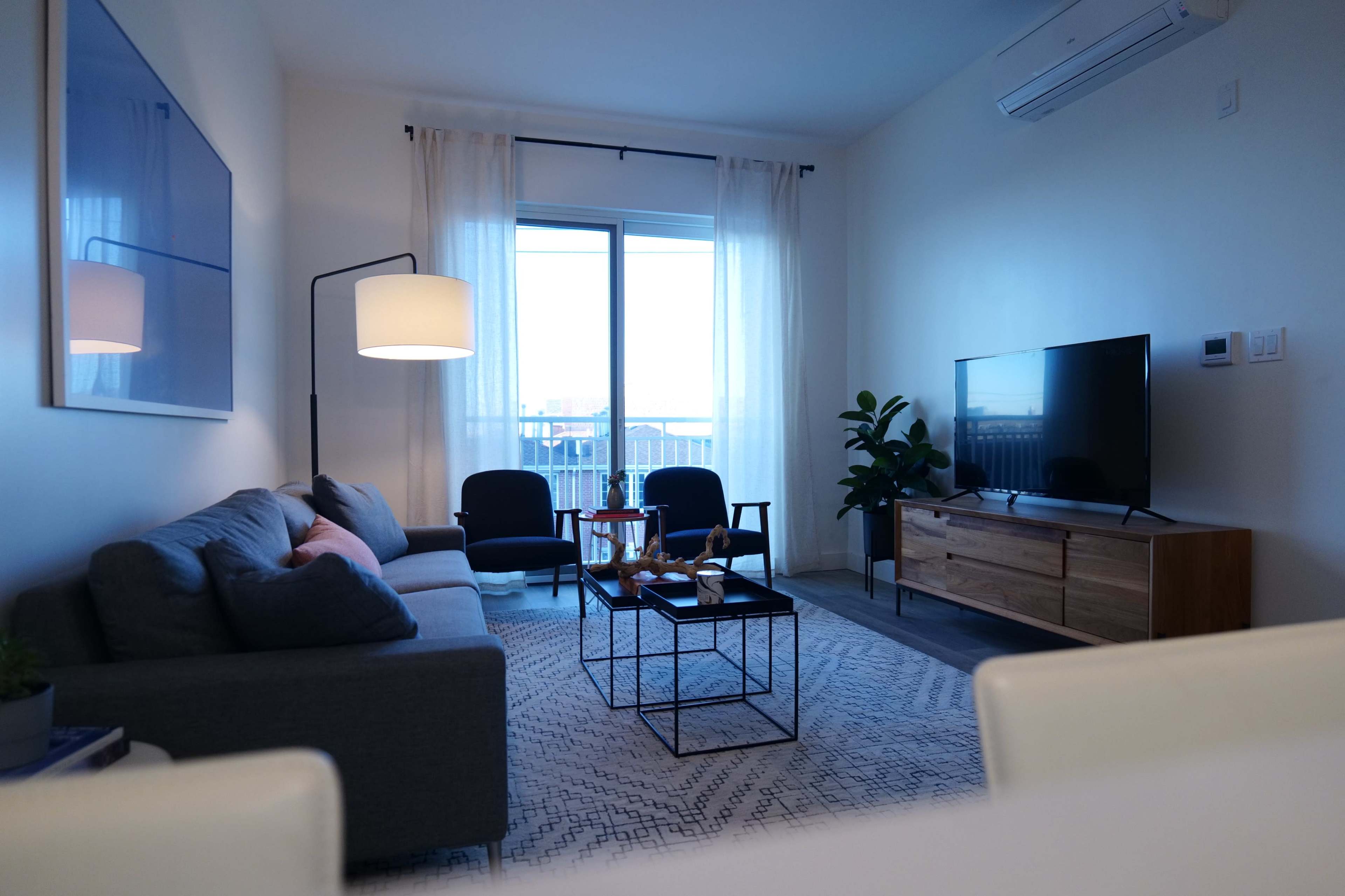 The living room features a gray sofa, two black chairs, a wooden TV stand, and a large window with sheer curtains.