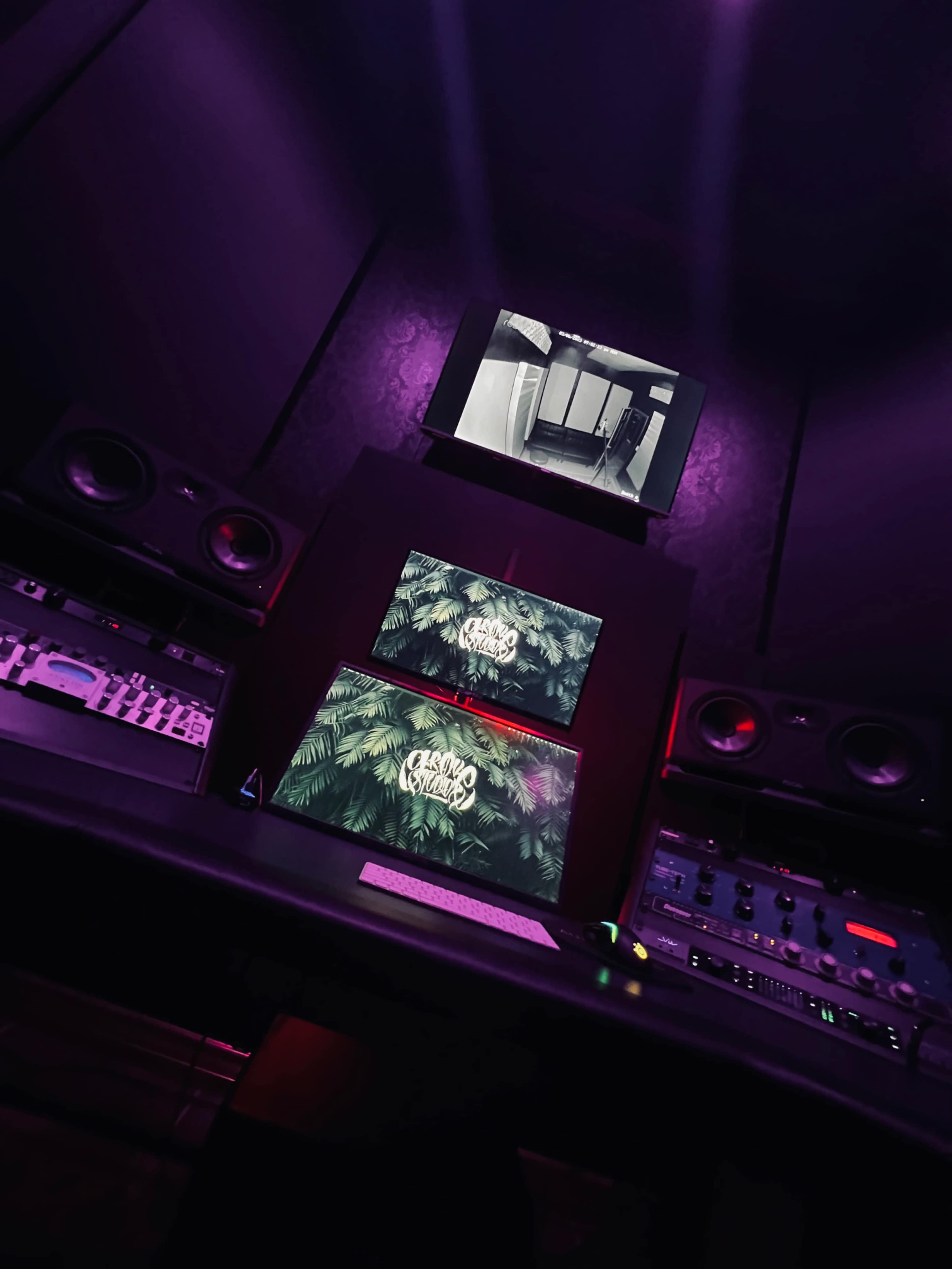 The image shows a music studio setup with two monitors displaying graphics, surrounded by audio equipment and speakers in a dimly lit environment.