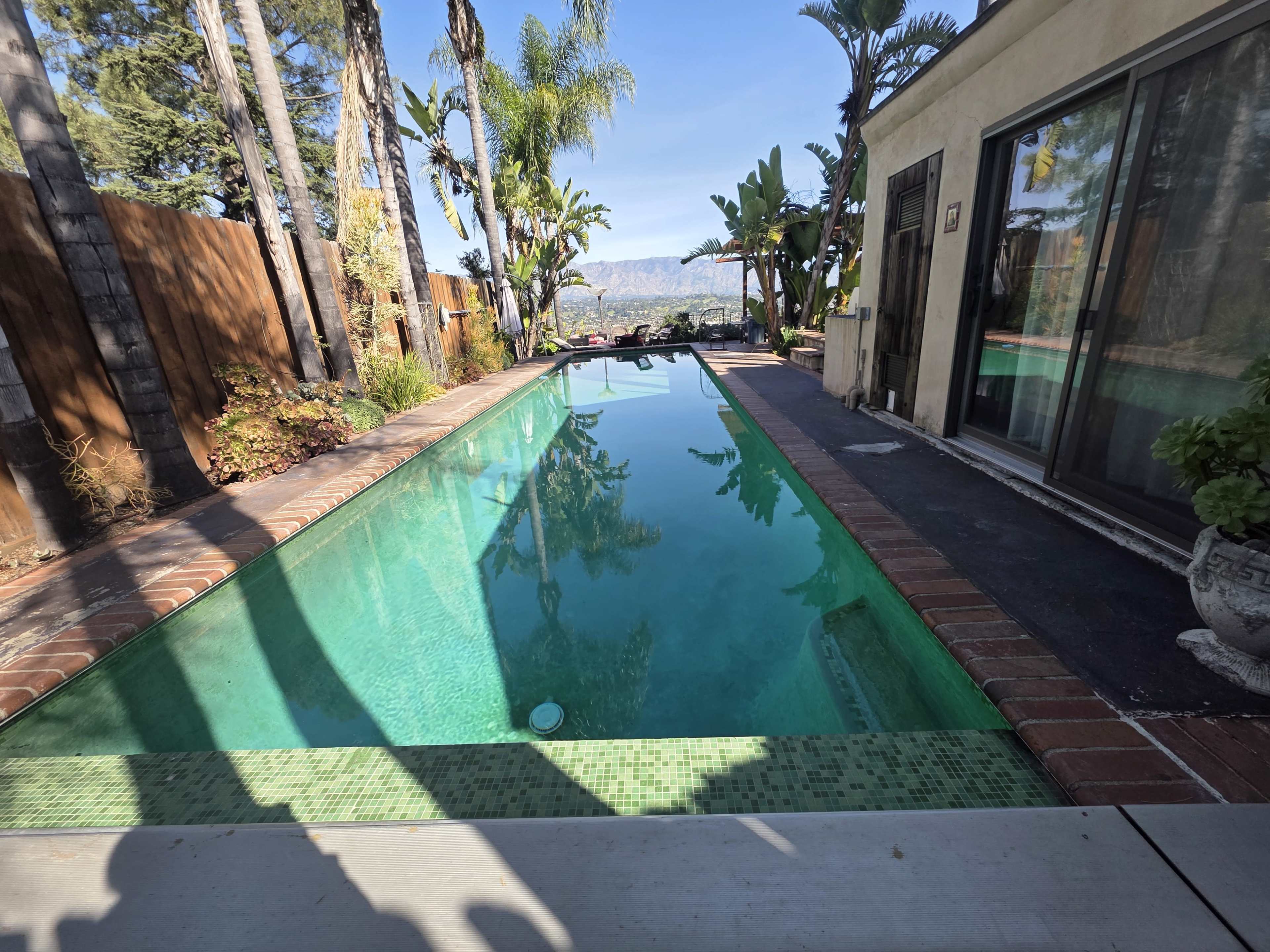 The image depicts a clear swimming pool surrounded by tropical plants and trees, with mountain views in the background.