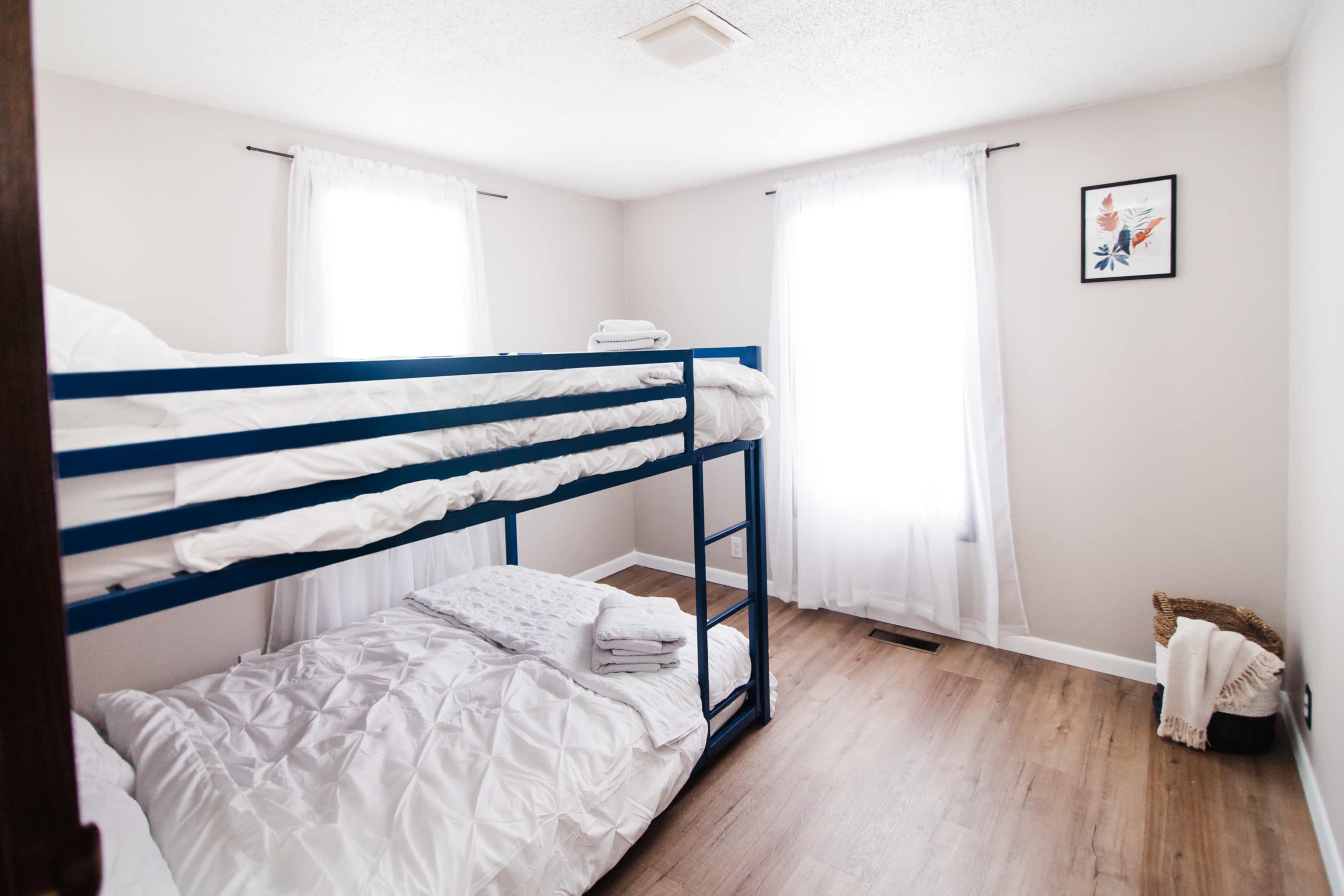 A simple bedroom featuring a bunk bed with white bedding, a single mattress on the floor, and two windows with sheer curtains.