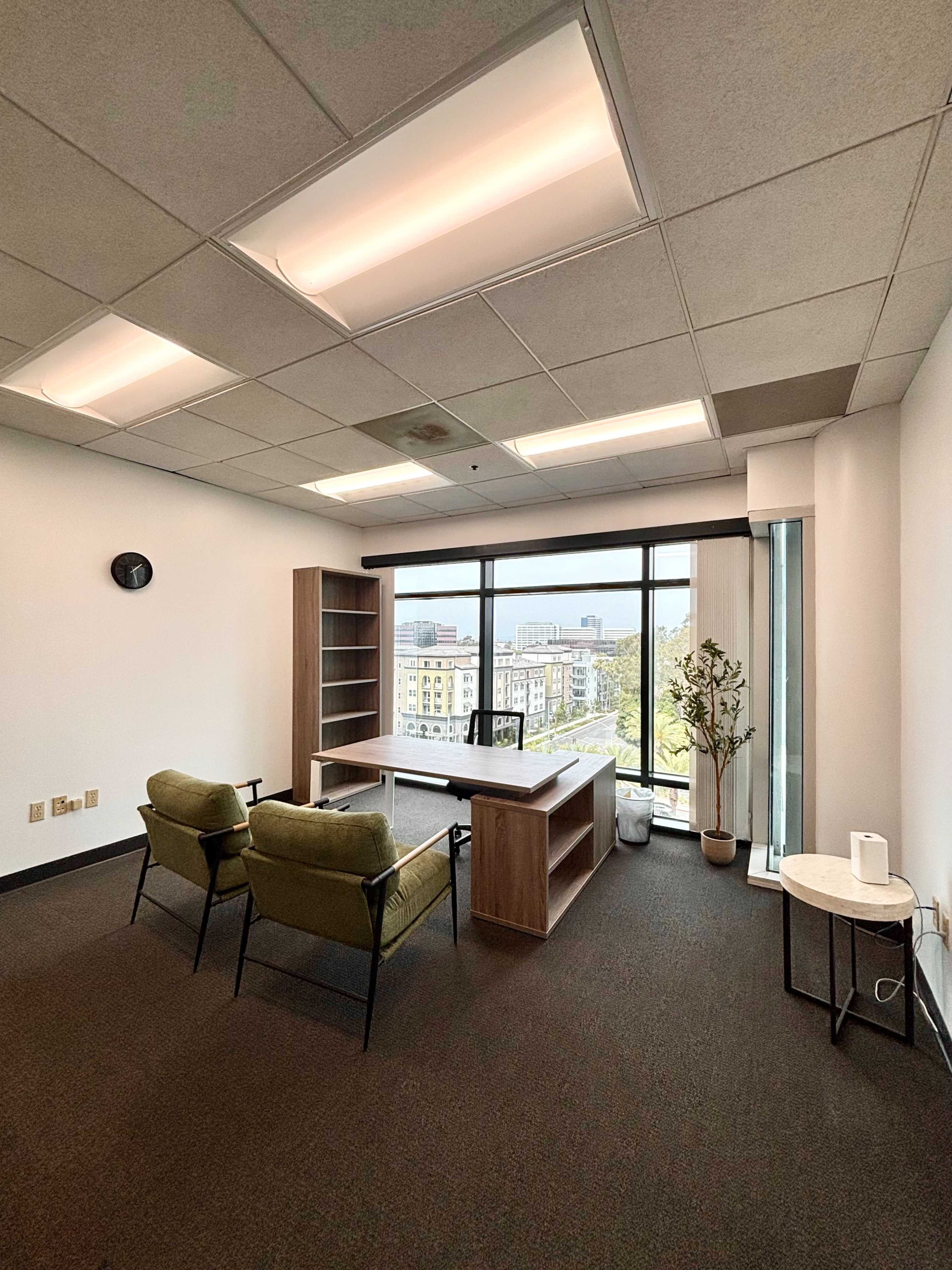 An empty office space features a desk, two chairs, a bookshelf, and a view of the cityscape through large windows.