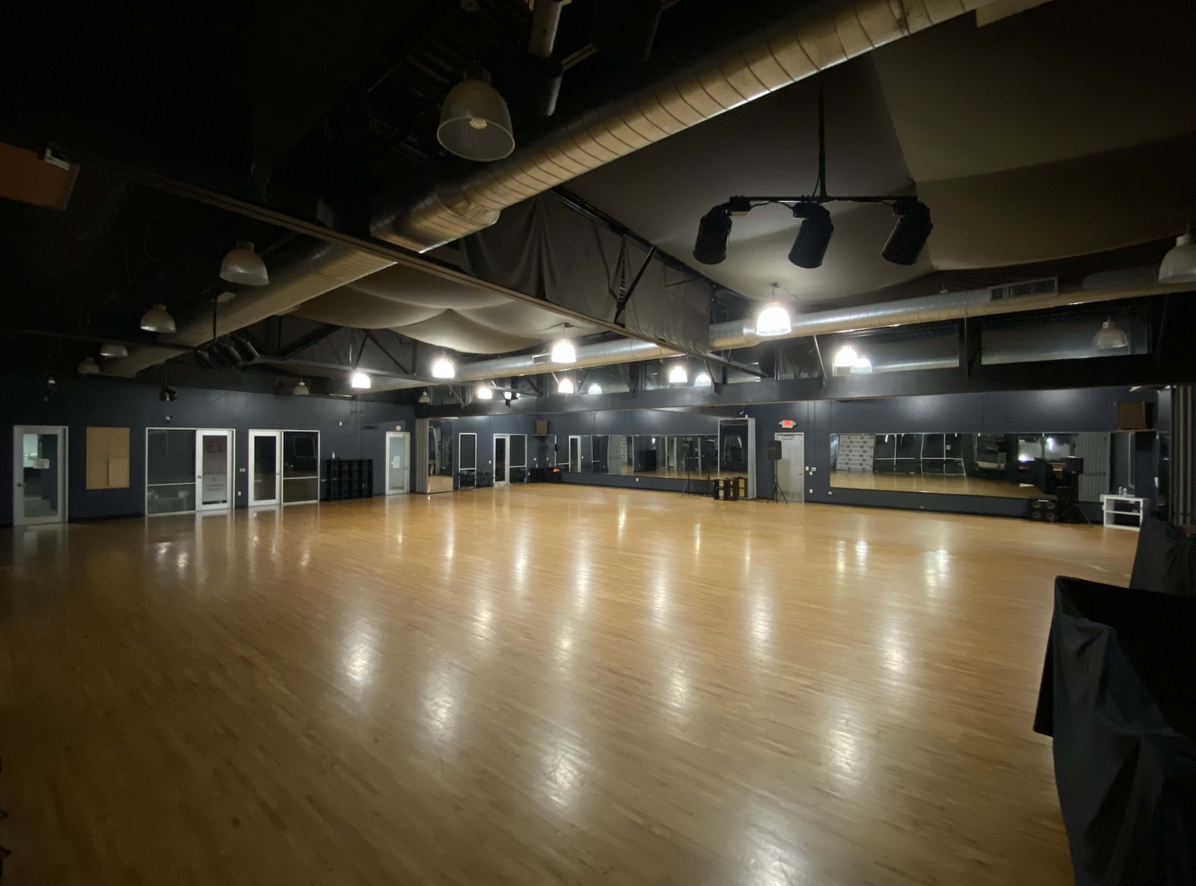 A large, empty dance studio with wooden flooring, mirrors on one wall, and overhead lighting in a dark-colored space.