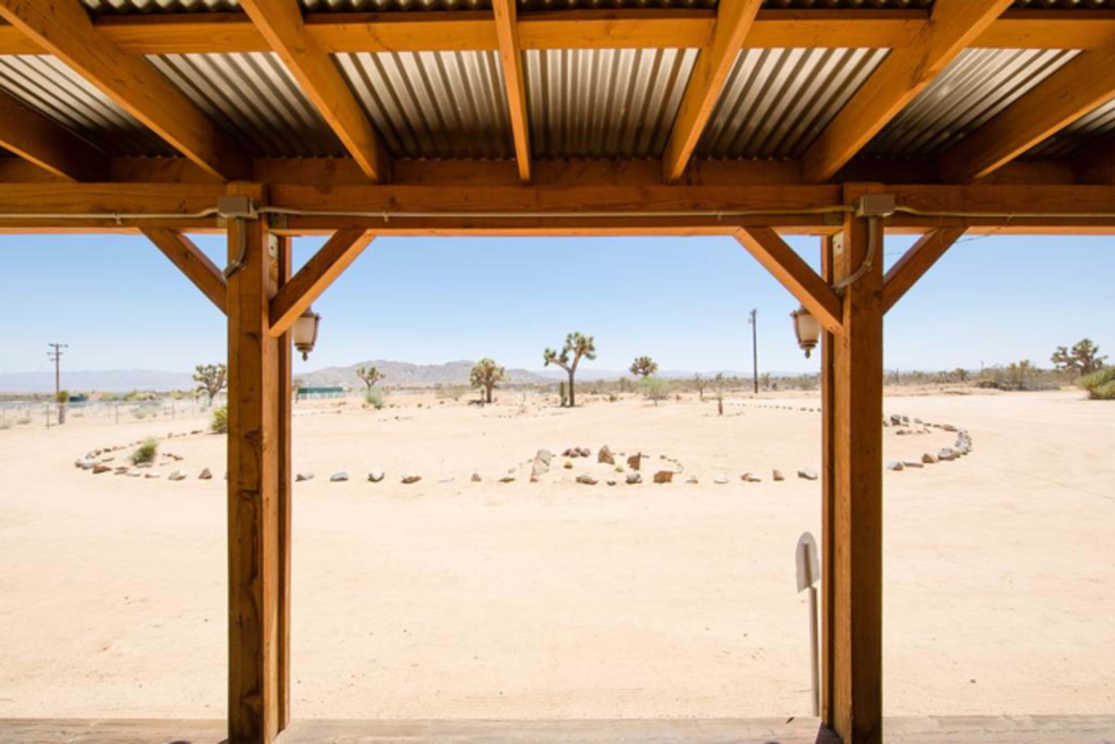 The image shows a wooden porch overlooking a barren landscape with a rock formation in a circular pattern and several Joshua trees in the distance.